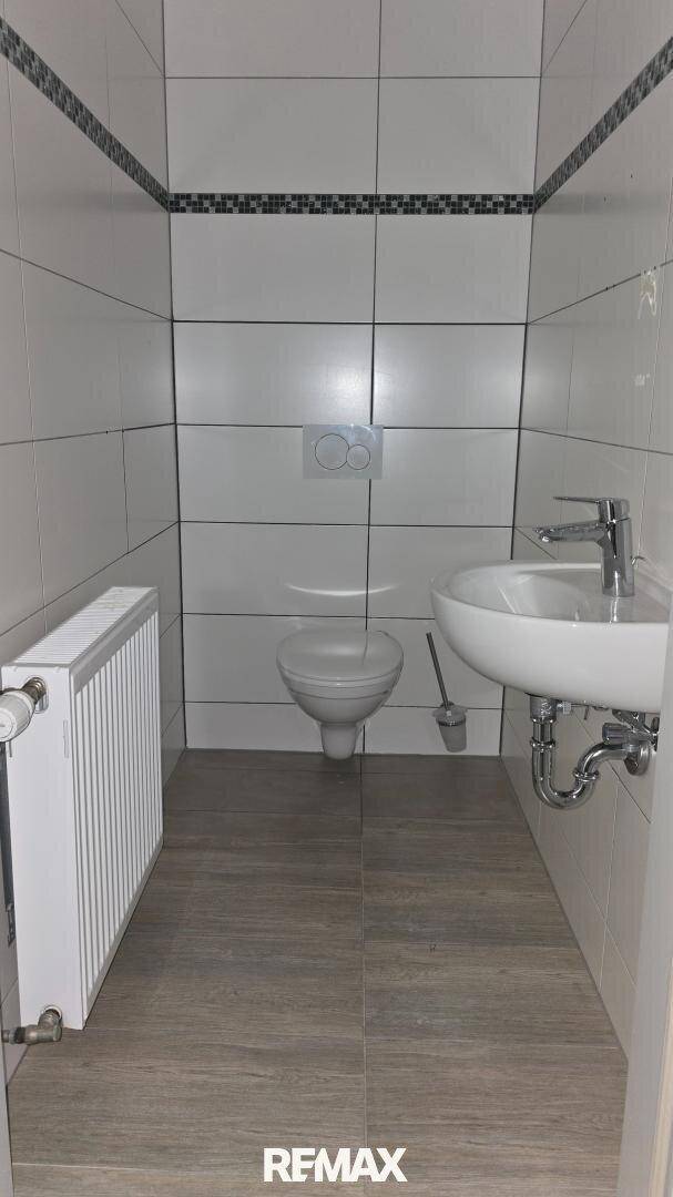 Modernes Gäste-WC mit wandhängender Toilette, Waschbecken und Heizkörper, gefliest in Weiß und Grau.