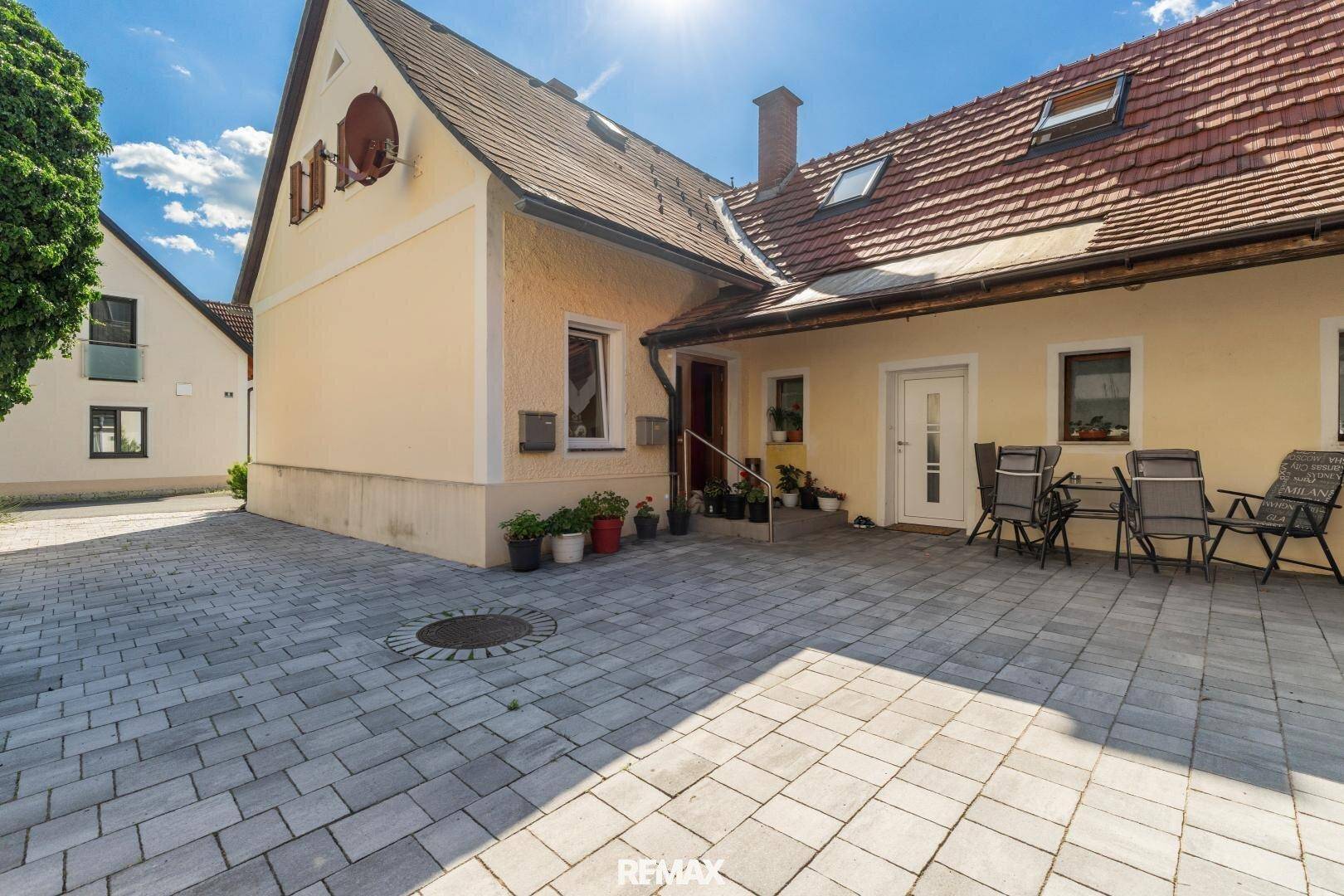 Traditionelles Gebäude mit gepflastertem Vorplatz, rotem Ziegeldach und blühenden Pflanzen.