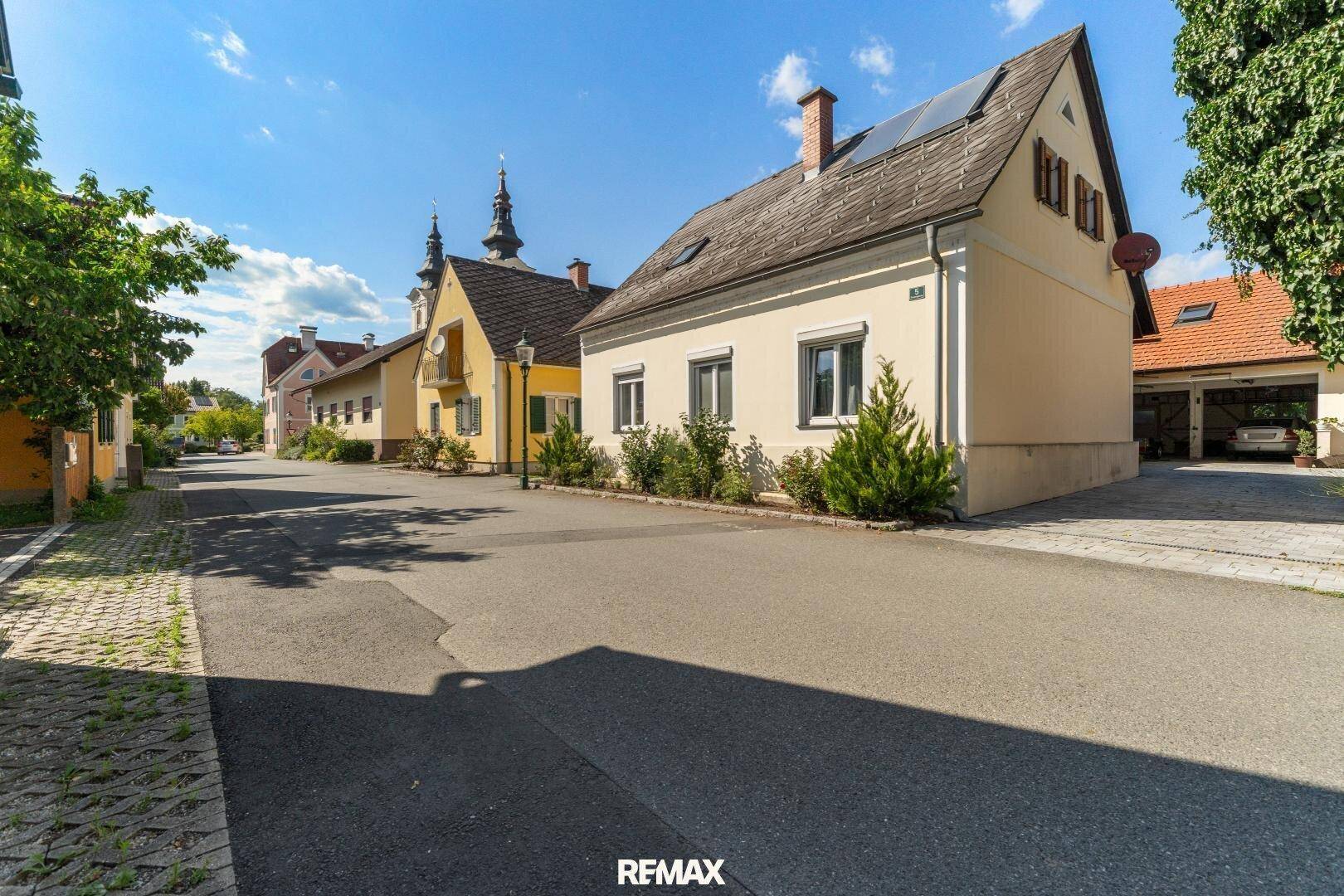 Ansicht des Gebäudes von der Straße mit umliegenden Häusern und einer Kirche im Hintergrund.