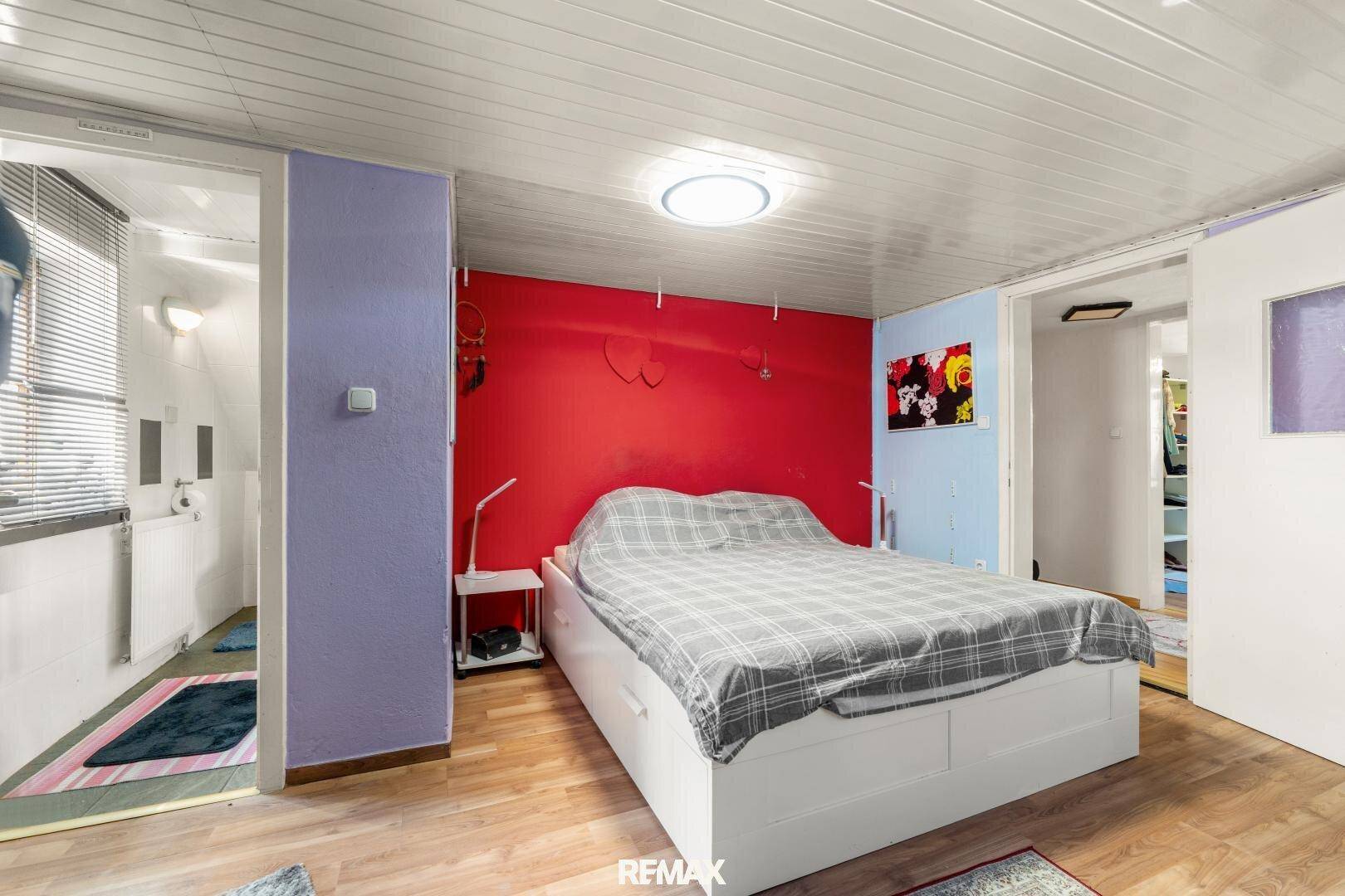Schlafzimmer mit roter Akzentwand, Holzboden und direktem Zugang zu einem kleinen Badezimmer.