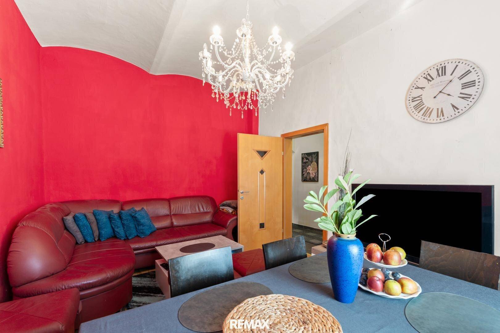 Gemütlicher Wohnbereich mit roter Akzentwand, Ledersofa und großer Wanduhr über dem Fernseher.