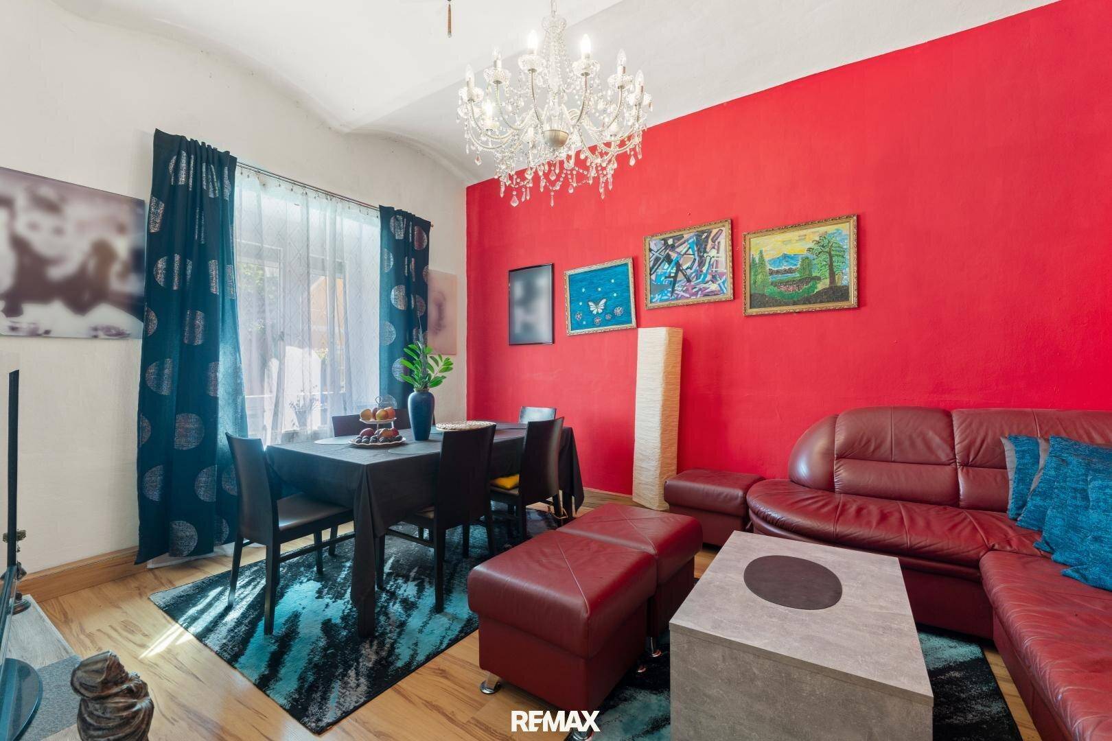 Farbenfroher Wohn-Essbereich mit roter Akzentwand, Ledersofa und elegantem Kronleuchter.