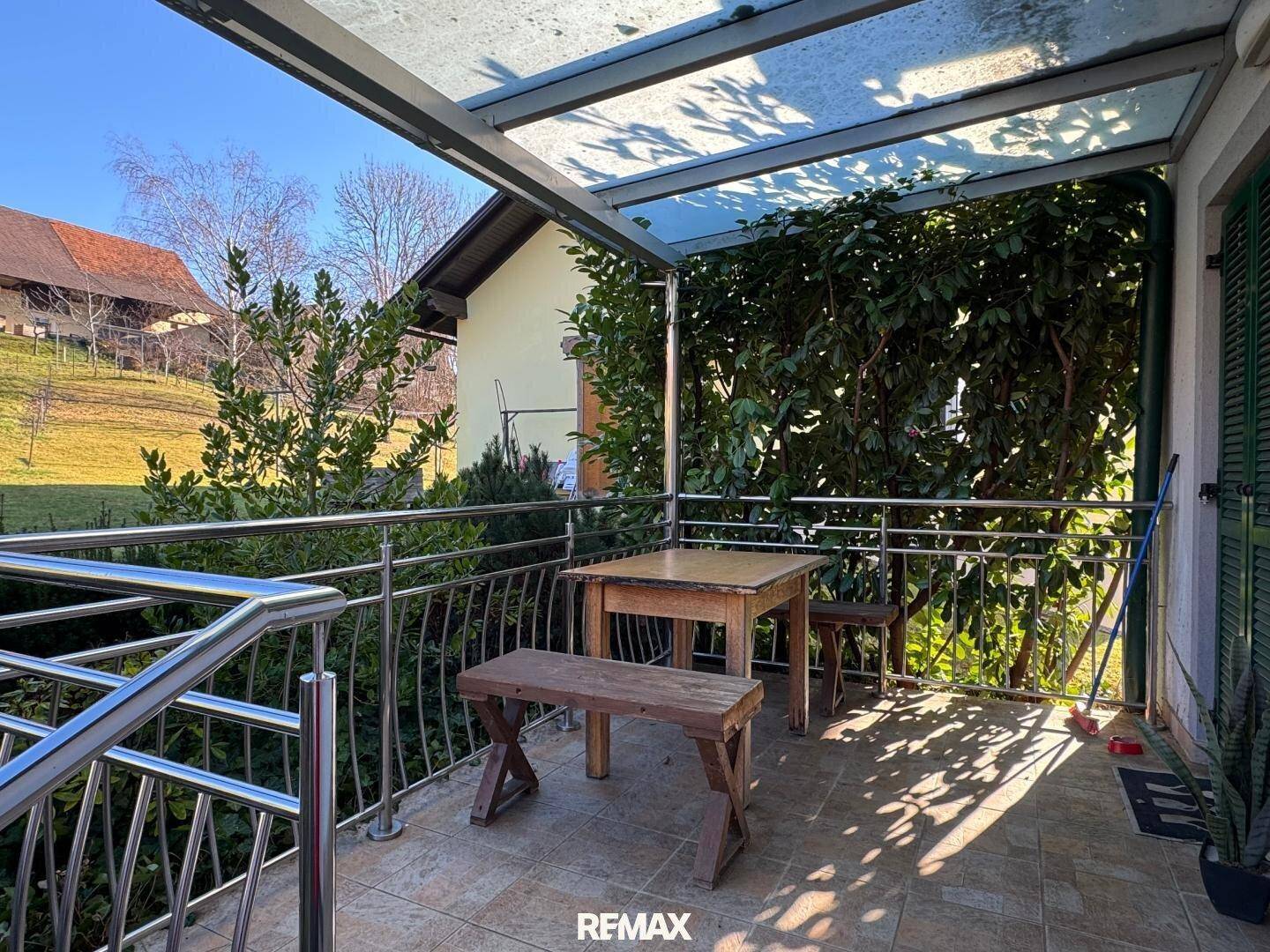 Überdachter Balkon mit Holztisch und Bänken, umgeben von üppigem Grün und Blick ins Grüne.