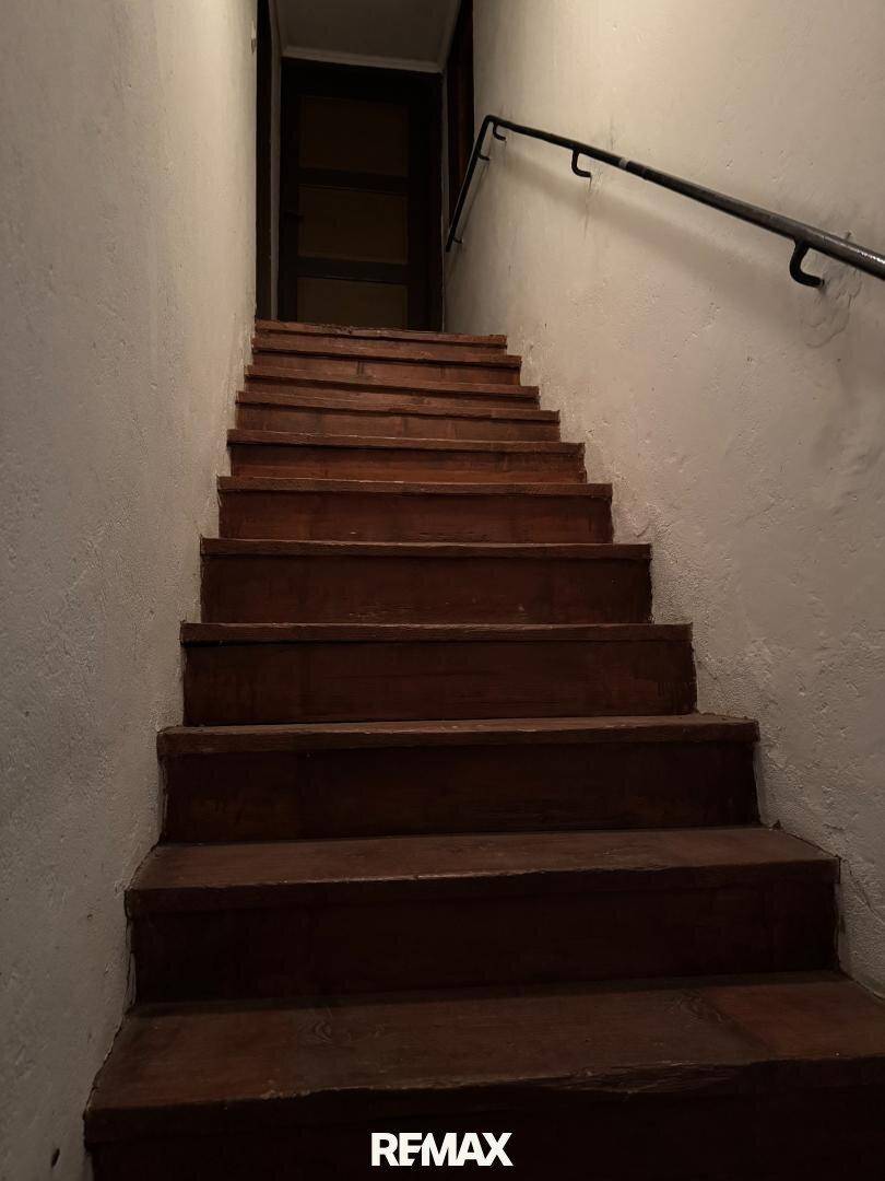 Dunkle Holztreppe mit Handlauf in einem schlichten Flur, der nach oben führt.