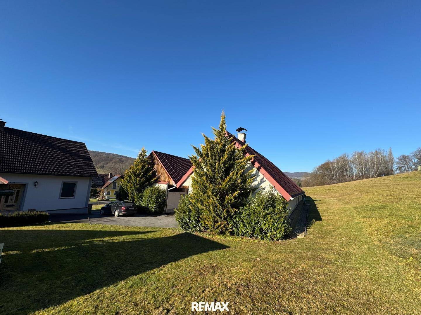Haus mit rotem Dach, umgeben von grünen Wiesen und Bäumen, unter strahlend blauem Himmel.