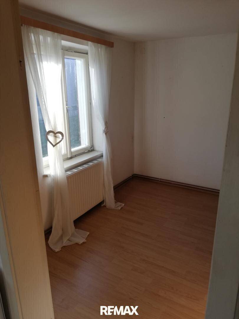 Gemütliches Zimmer mit Holzboden und einem Fenster, das durch weiße Vorhänge sanftes Licht filtert.