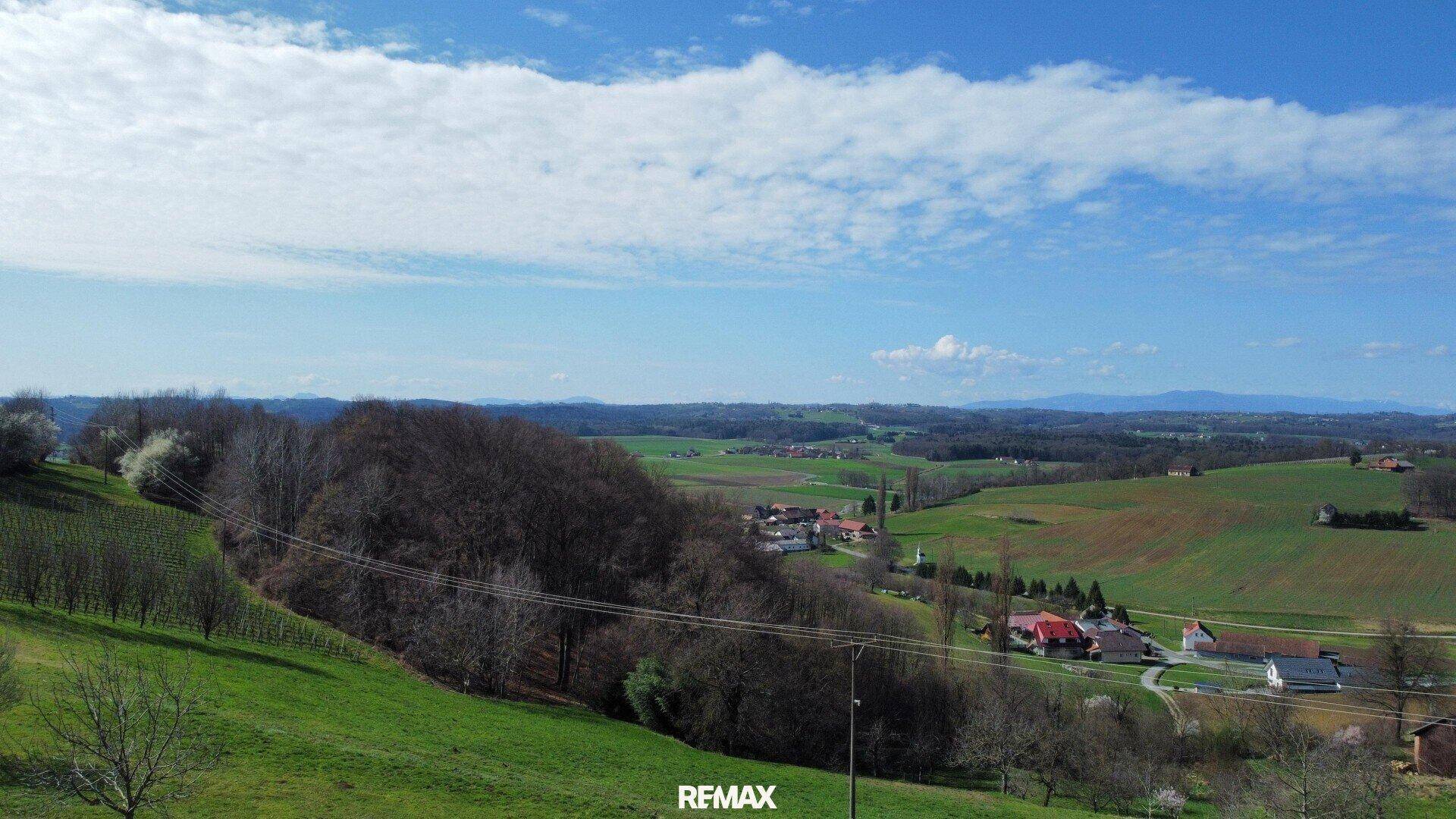 Weitläufige Landschaft mit grünen Hügeln, Feldern und einem Dorf unter blauem Himmel.