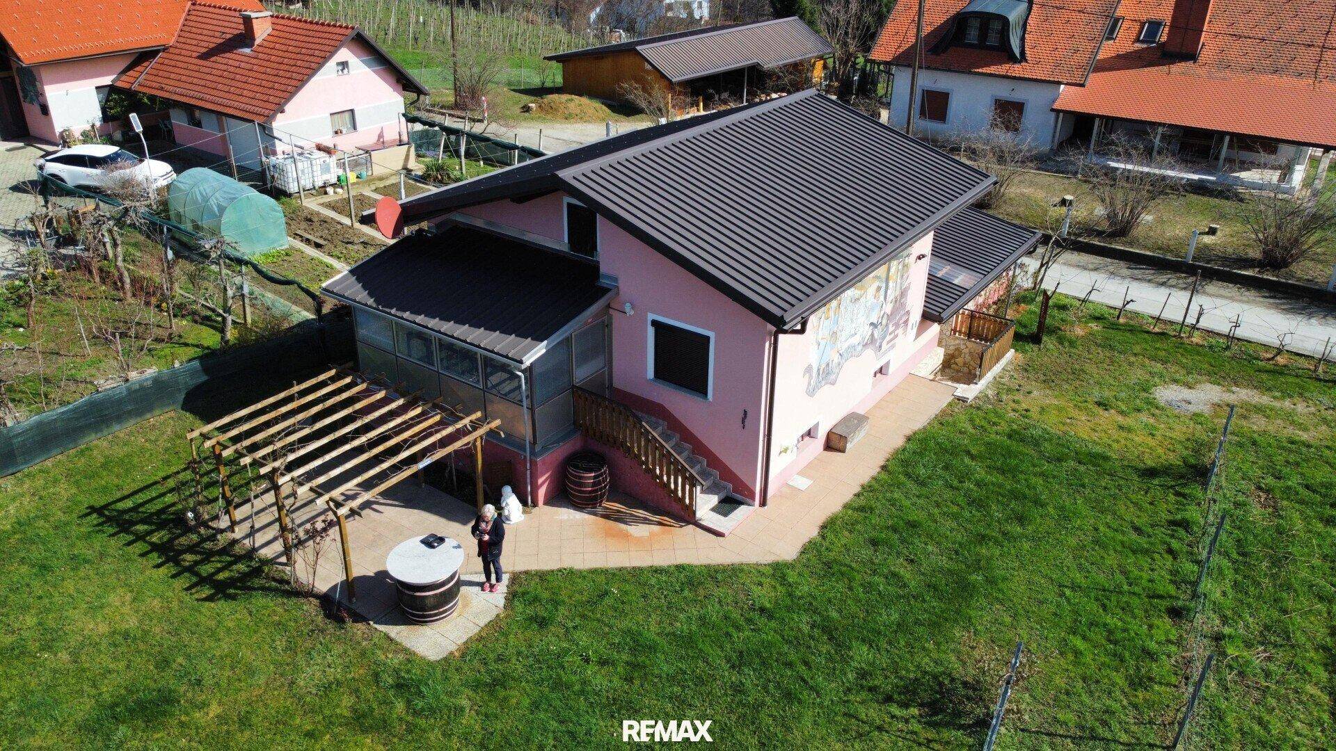Luftaufnahme eines rosa Hauses mit dunklem Dach, umgeben von Grünflächen und einer Pergola.