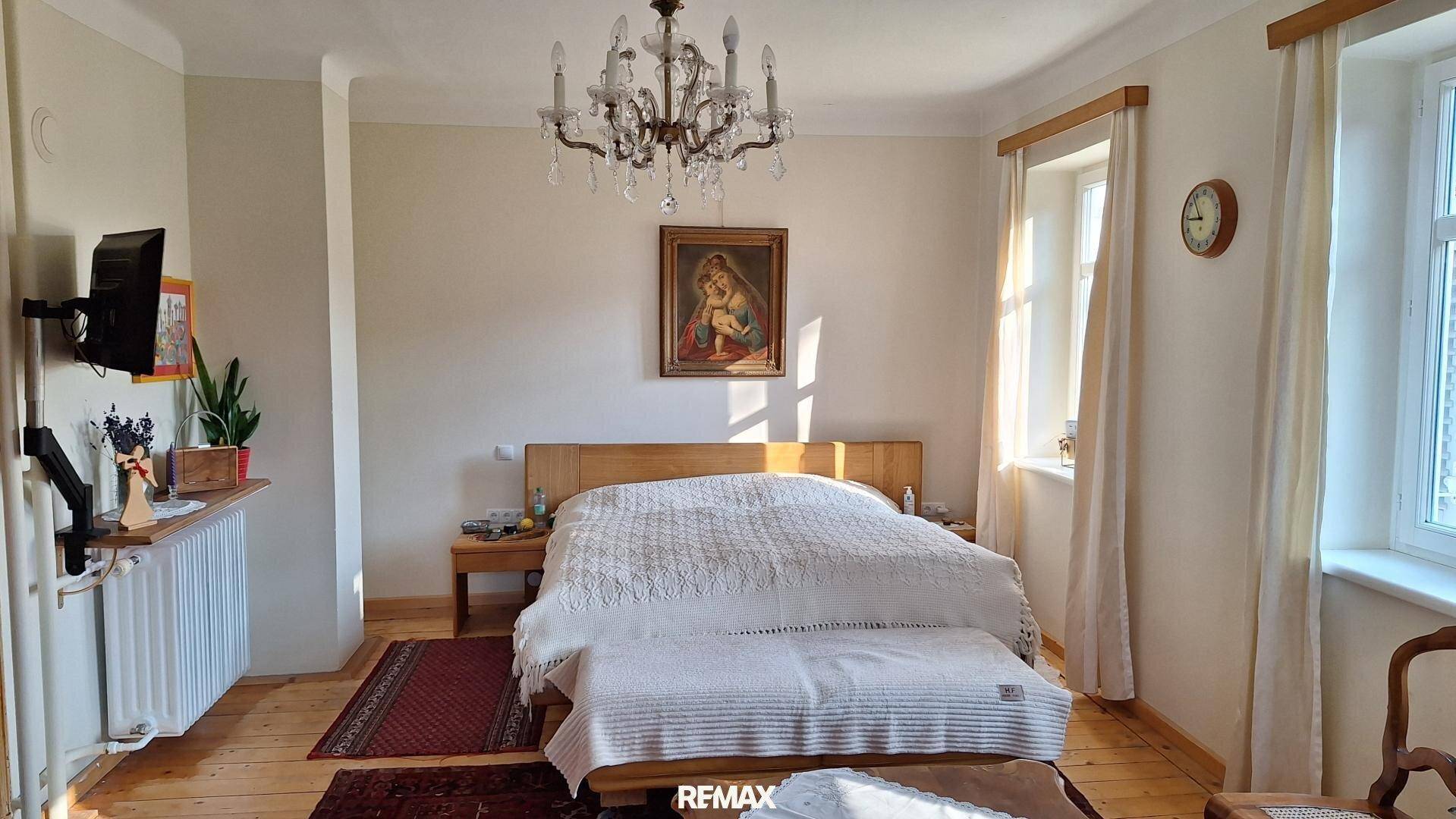 Helles Schlafzimmer mit Holzbett, Kronleuchter und Holzdielenboden, durchflutet von Tageslicht.