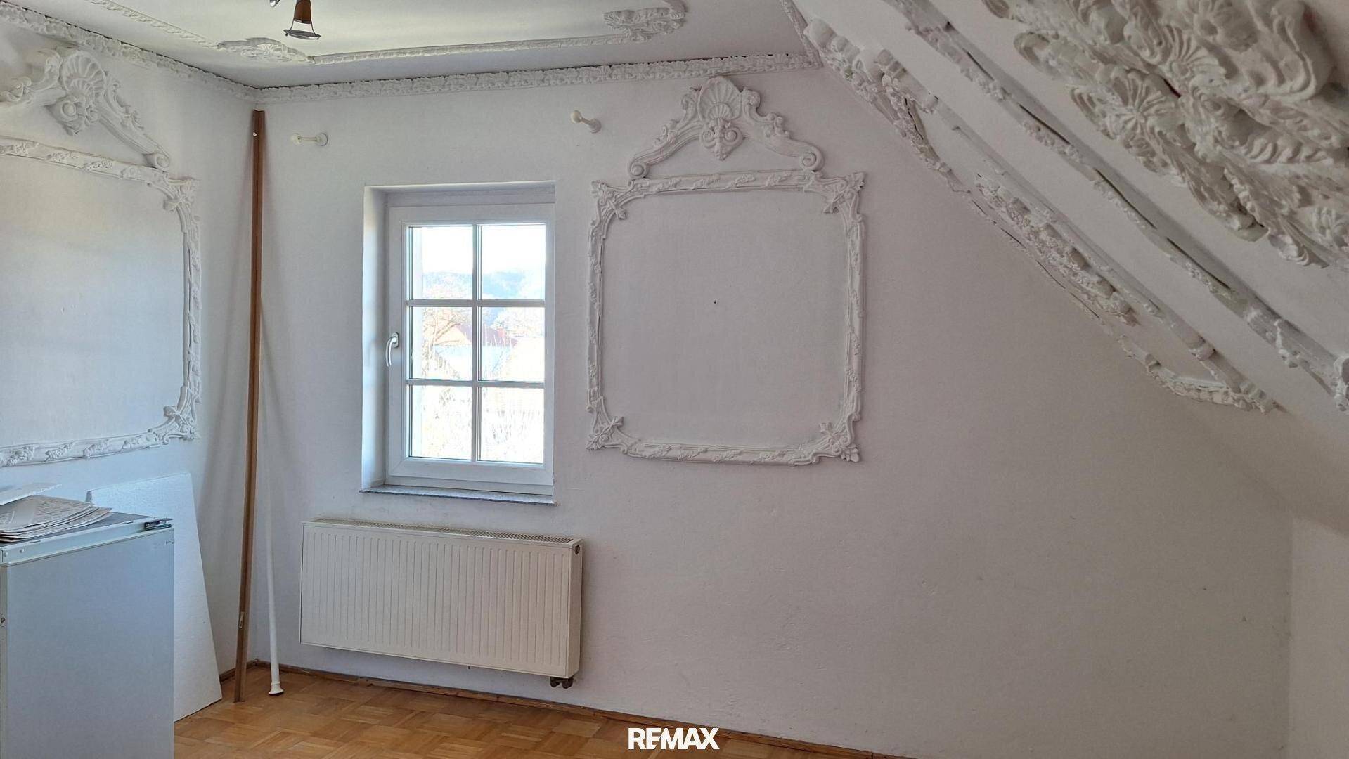 Dachzimmer mit aufwendiger Stuckverzierung an Wänden und Decke, Fenster und Heizkörper.