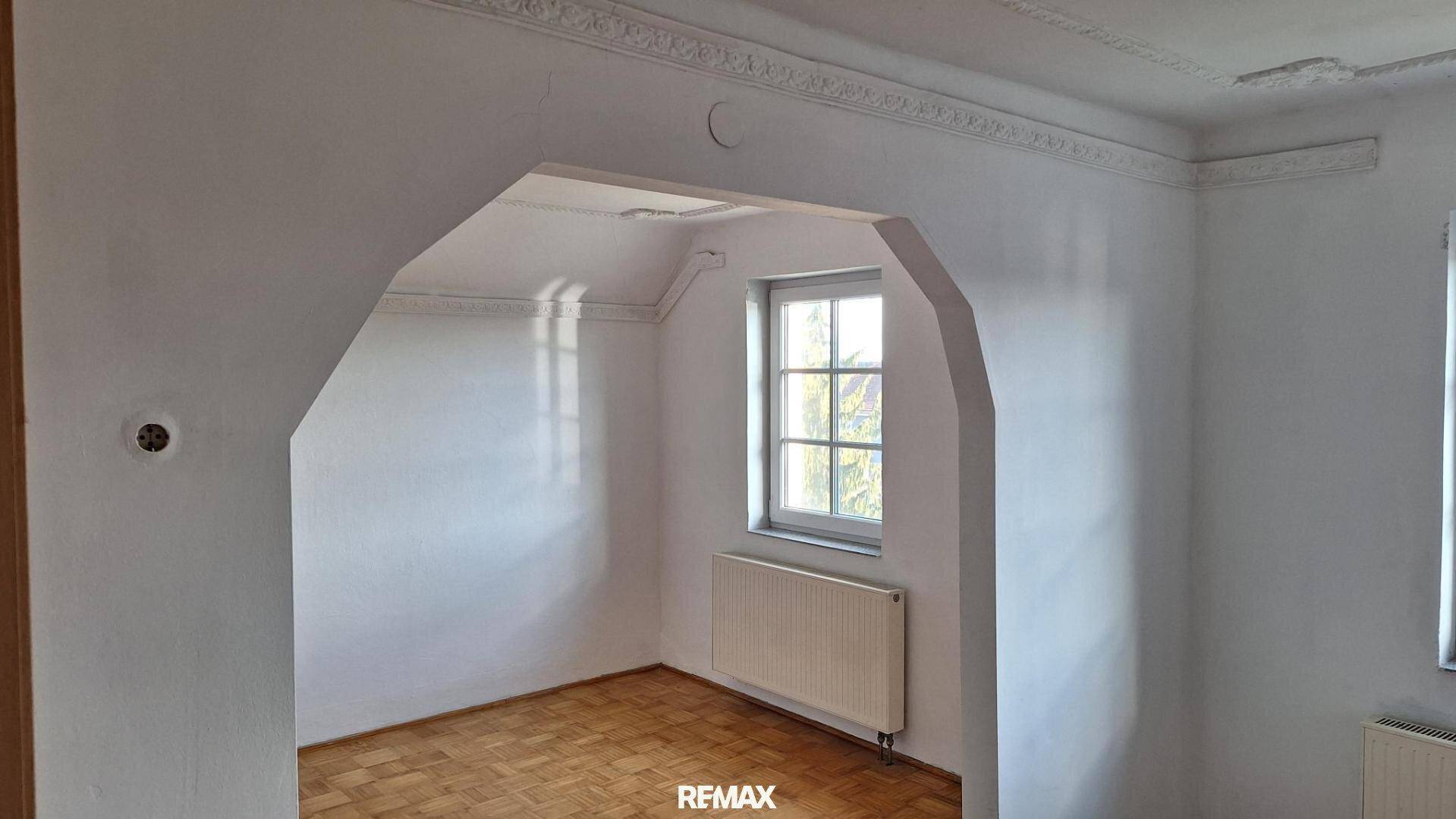 Helles Zimmer mit Parkettboden, Fenster, Heizkörper und einem dekorativen Bogen.
