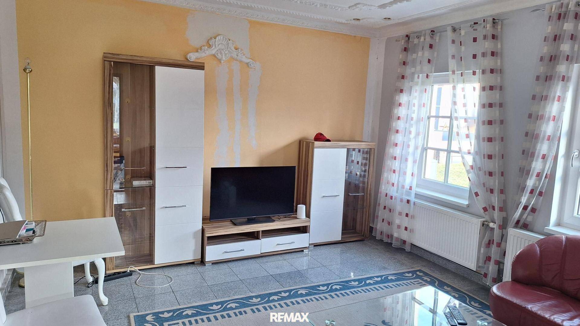 Wohnzimmer mit TV-Möbeln, kleinem Esstisch, rotem Sessel und Fliesenboden.