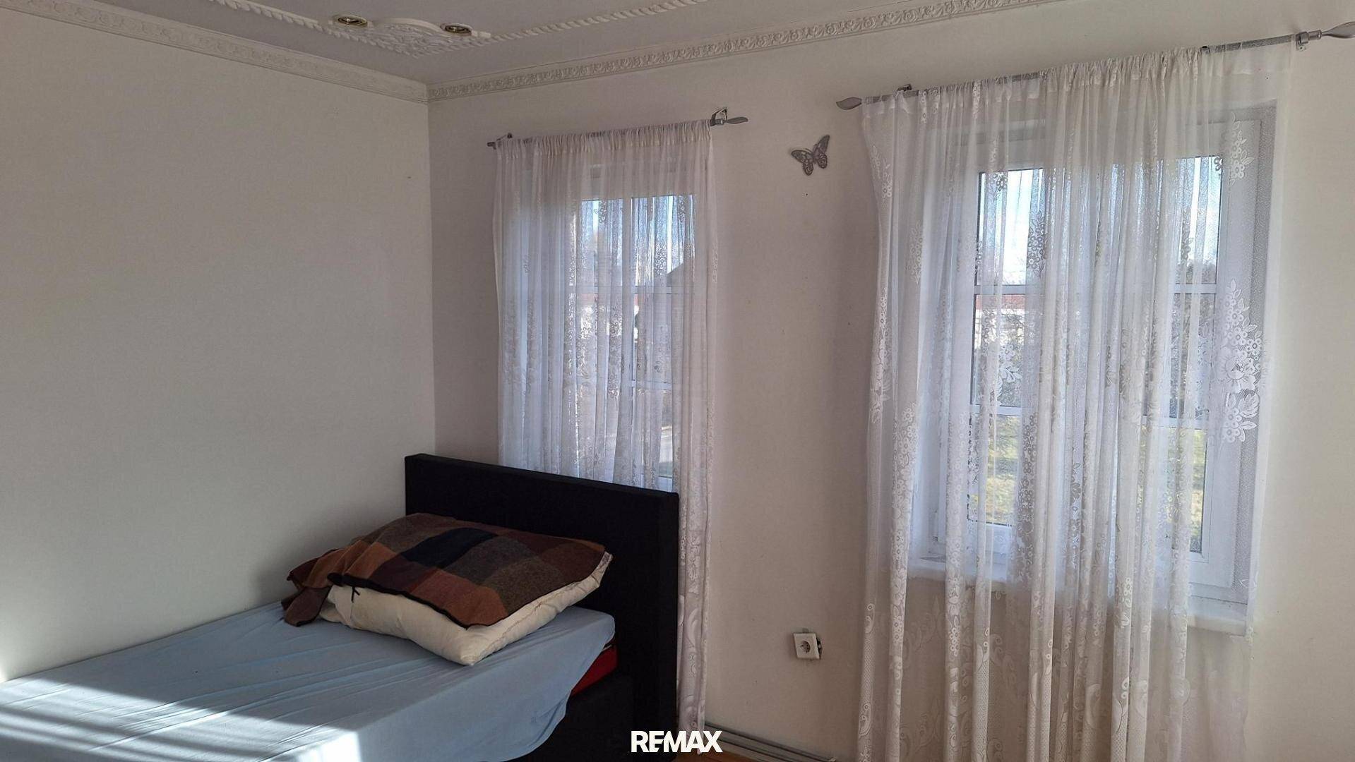 Schlafzimmer mit Einzelbett, zwei Fenstern mit Vorhängen und weiß gestrichenen Wänden.