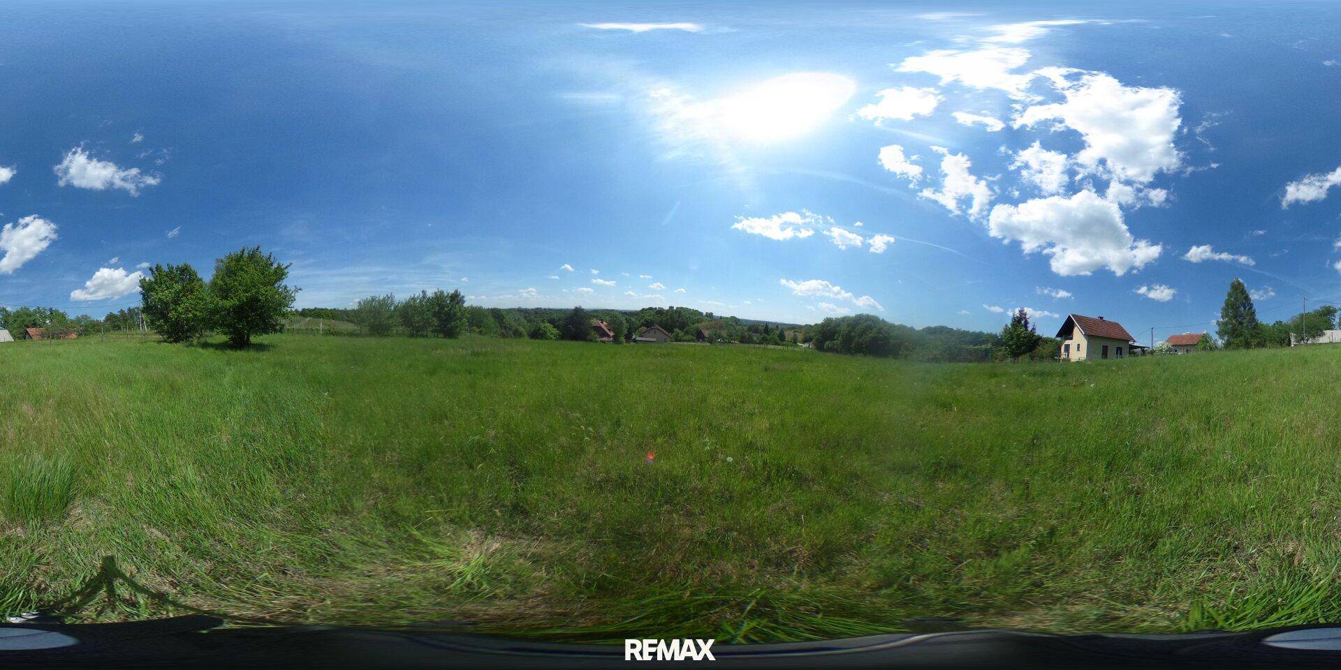 Panoramablick über ein weites grünes Feld mit Häusern und Bäumen am Horizont unter blauem Himmel.