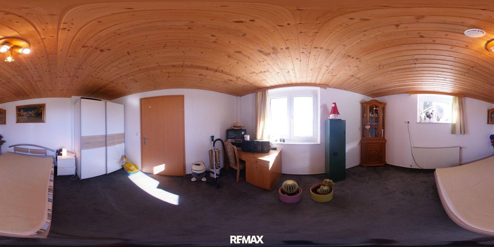 Schlafzimmer mit Holzbalkendecke, Teppichboden, Bett und Schreibtisch, hell durch die Fenster.