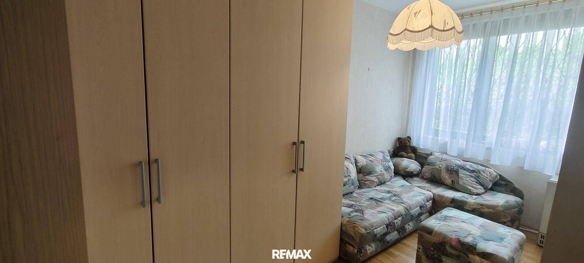 Helles Zimmer mit Sofa, Kleiderschrank und Fenster, das viel Tageslicht hereinlässt.