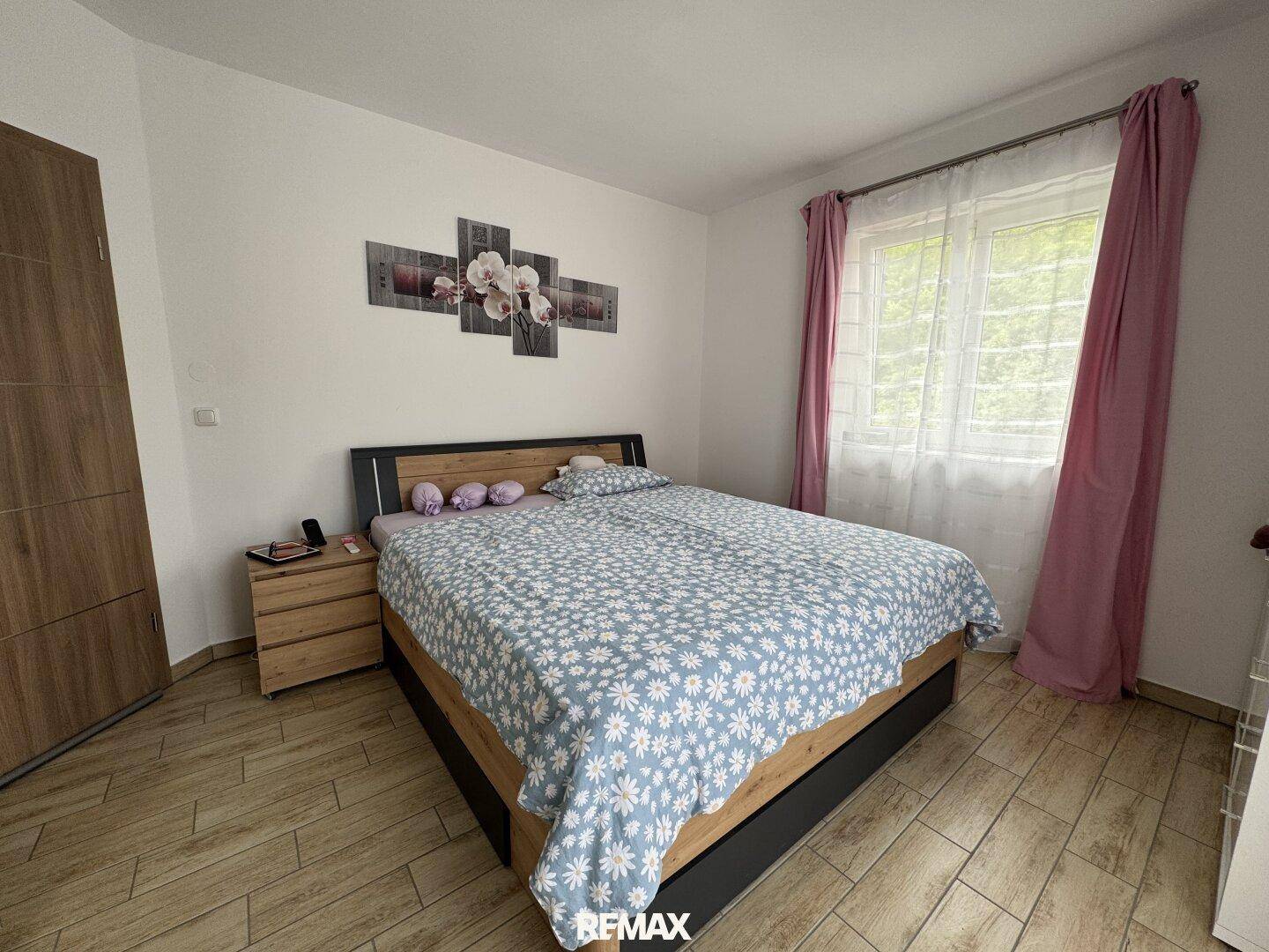 Gemütliches Schlafzimmer mit Doppelbett, Laminatboden und einem Fenster mit Blick ins Grüne.