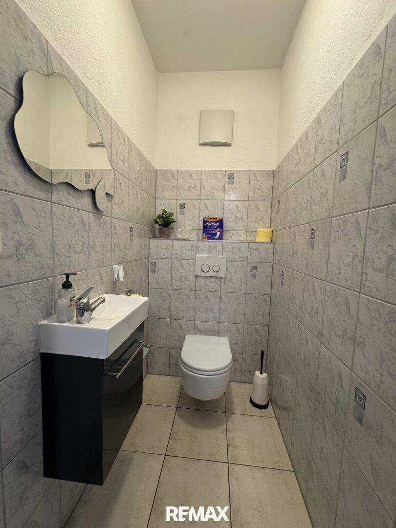 Modernes Gäste-WC mit Waschbecken, Wand-WC und einem Spiegel in ungewöhnlicher Form.