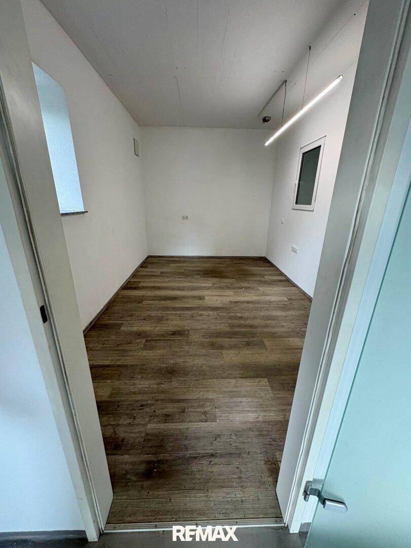 Kleiner, heller Raum mit Laminatboden, weißen Wänden und zwei Fenstern, durch eine offene Tür gesehen.