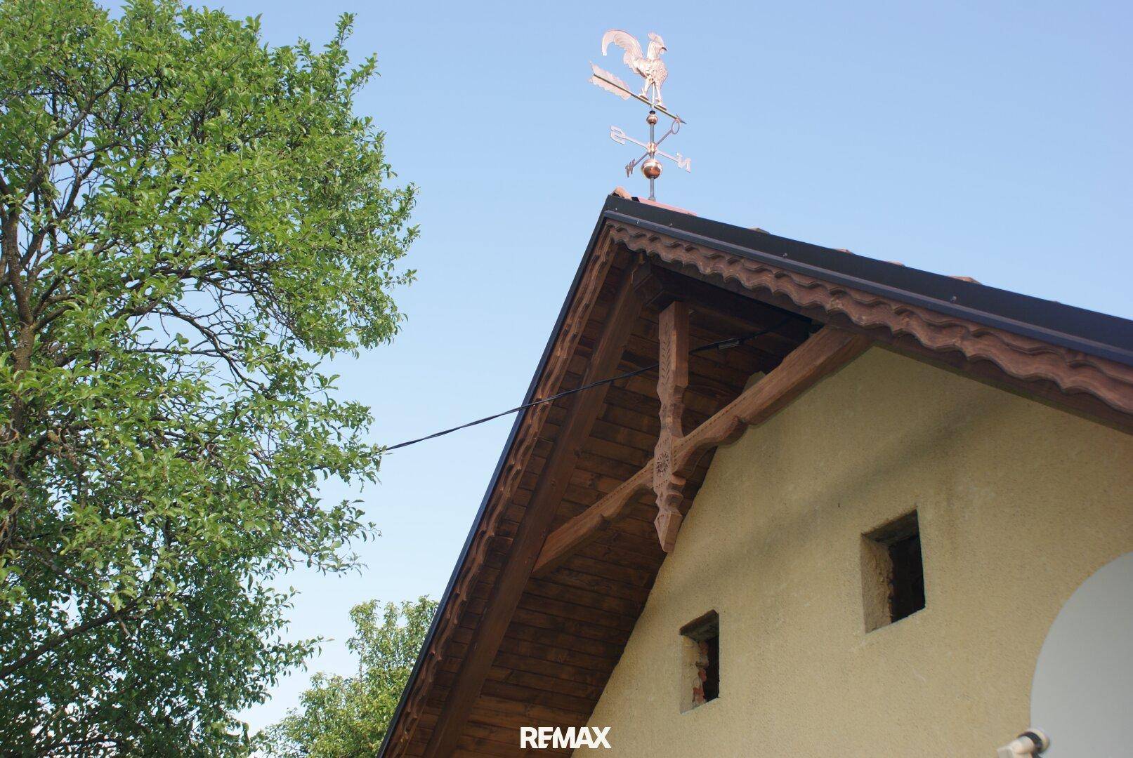 Detailansicht des Daches mit Holzbalken und einem Wetterhahn unter blauem Himmel.