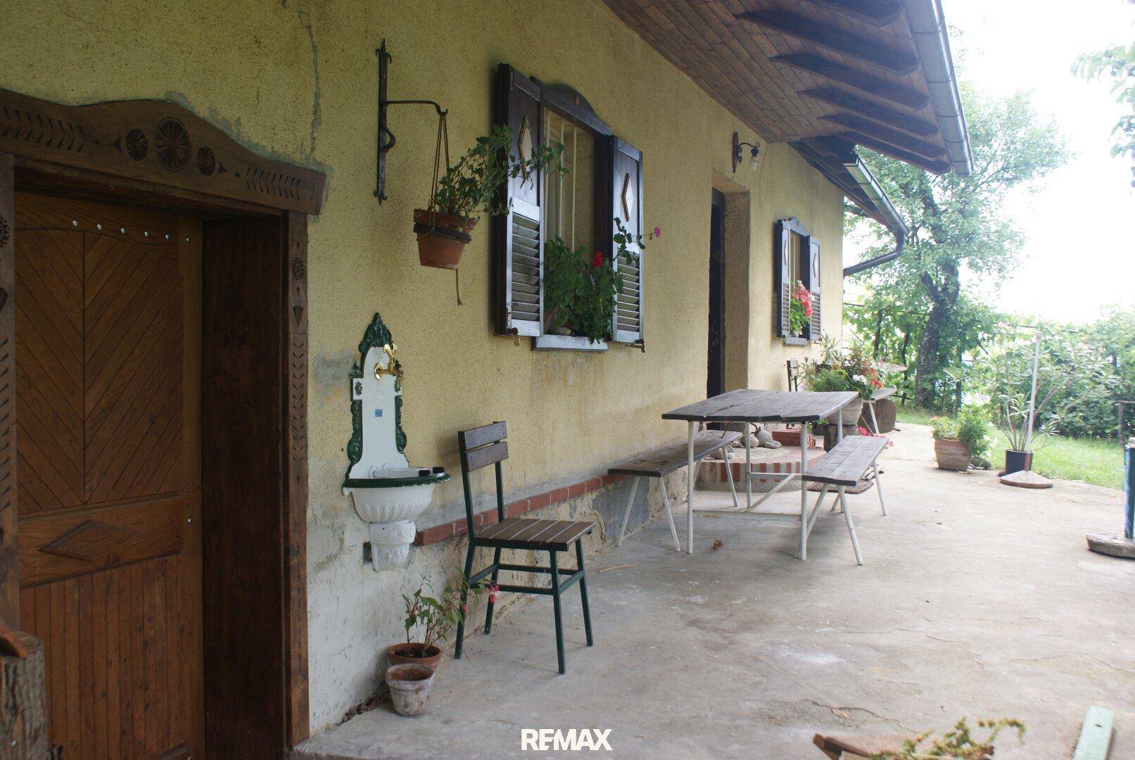 Rustikale Terrasse vor einem alten Haus mit Holztisch, Bänken und einem dekorativen Wandbrunnen.