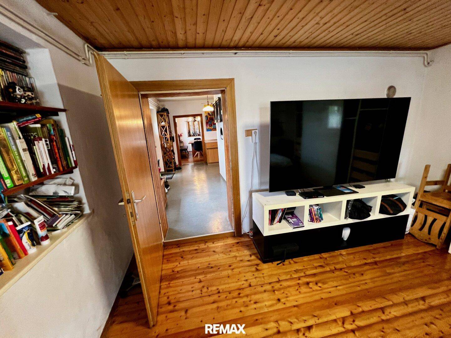 Schlafzimmer mit Holzboden und Blick in den Flur, ausgestattet mit einem großen Fernseher.