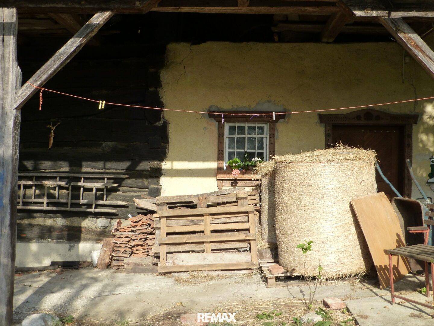 Detailansicht des überdachten Bereichs mit Holzpaletten und Heuballen vor der Hauswand.