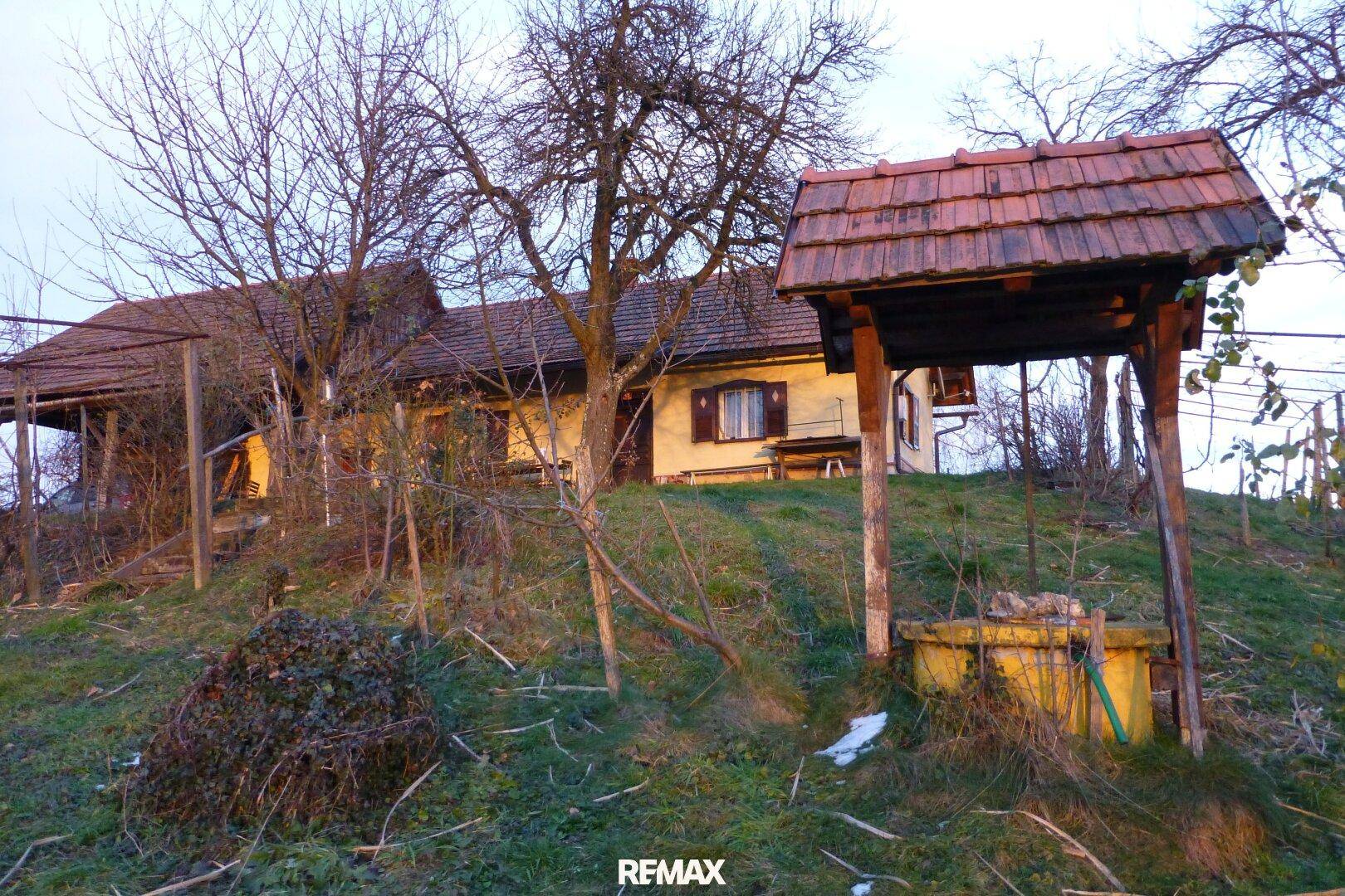 Traditionelles Haus auf einem Hügel mit einem alten Ziehbrunnen im Vordergrund und kahlen Bäumen.