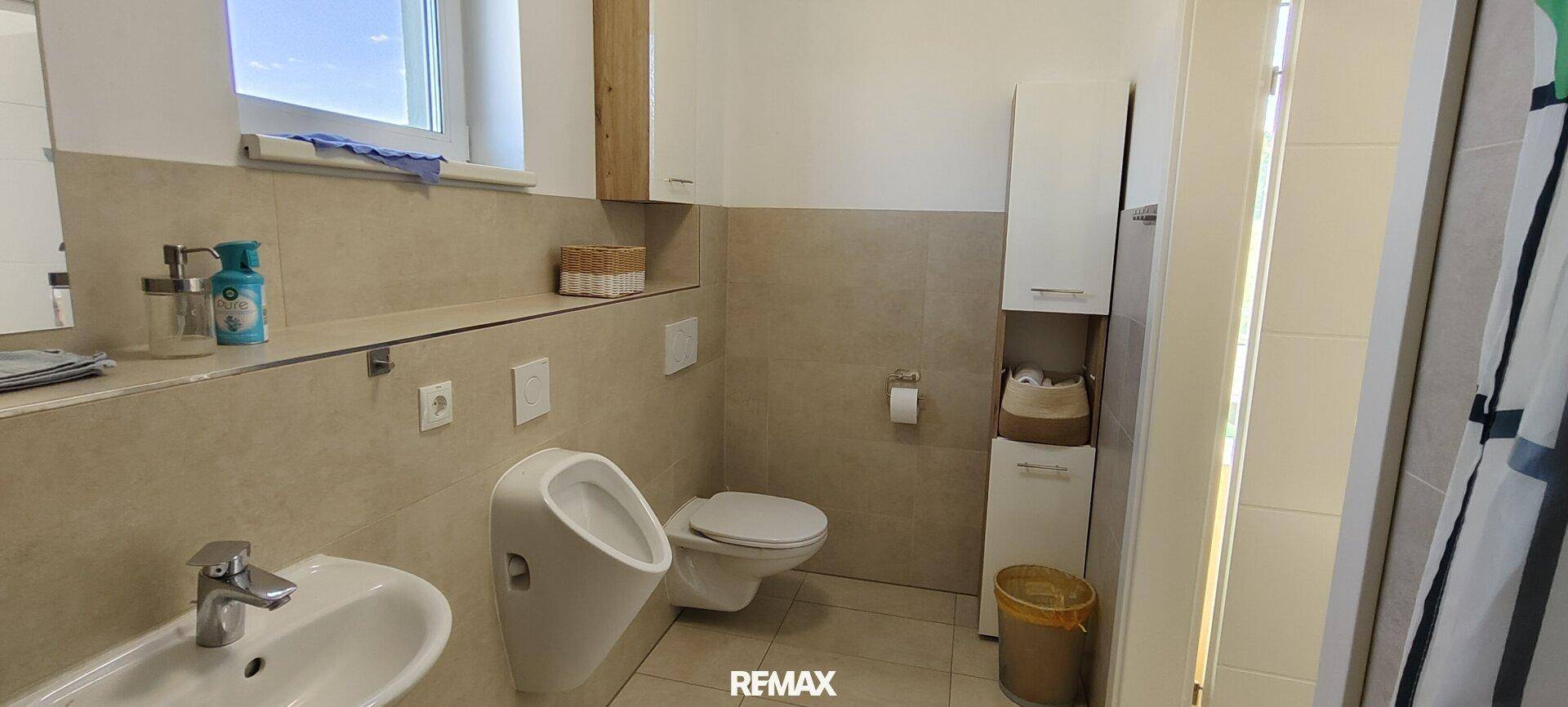 Modernes Badezimmer mit WC, Urinal, Waschbecken und Fenster, hell gefliest und funktional.