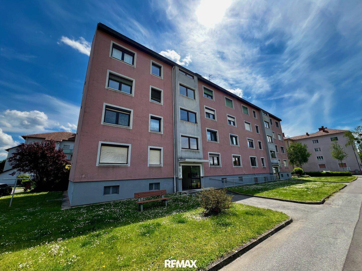 Mehrfamilienhaus mit rosafarbener Fassade und grüner Umgebung unter blauem Himmel.