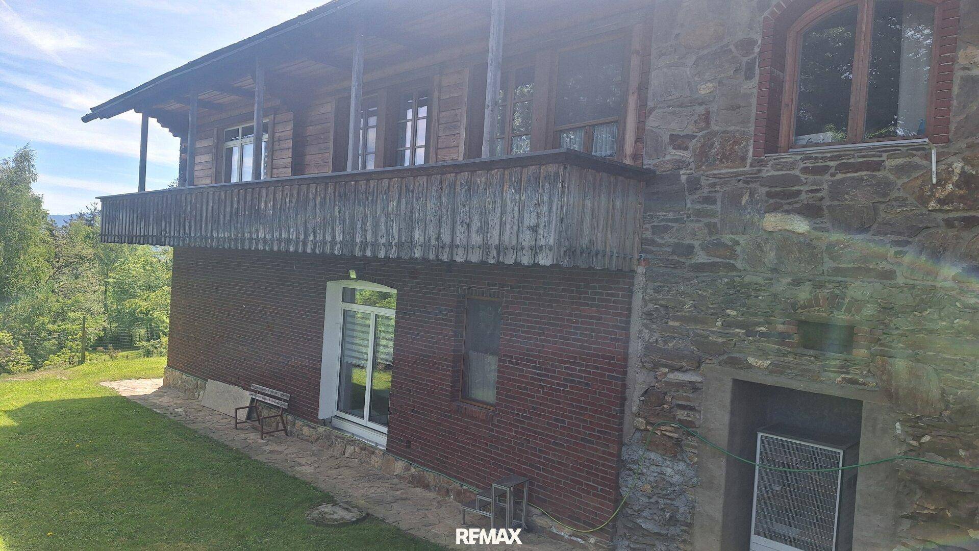 Seitenansicht des Hauses mit Holzbalkon, Ziegelmauerwerk und Steinfassade, umgeben von Grün.