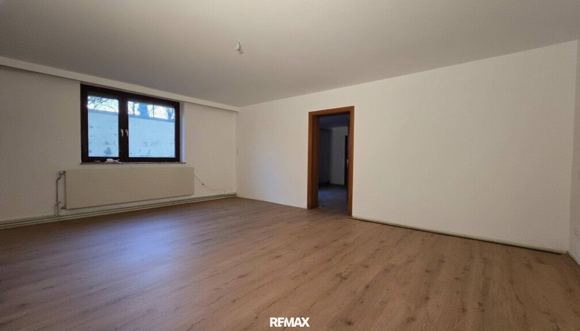 Heller, leerer Raum mit Laminatboden, weißen Wänden und einem Fenster mit Heizkörper.