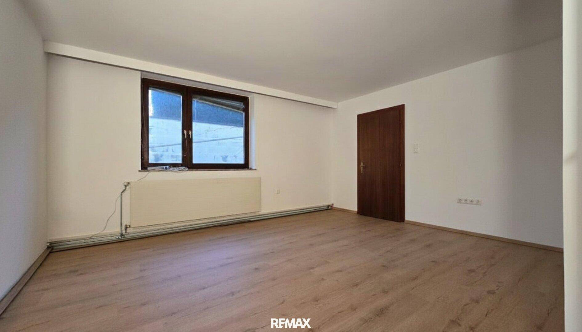 Geräumiger, unmöblierter Raum mit Laminatboden, weißen Wänden und einem Fenster mit Heizung.