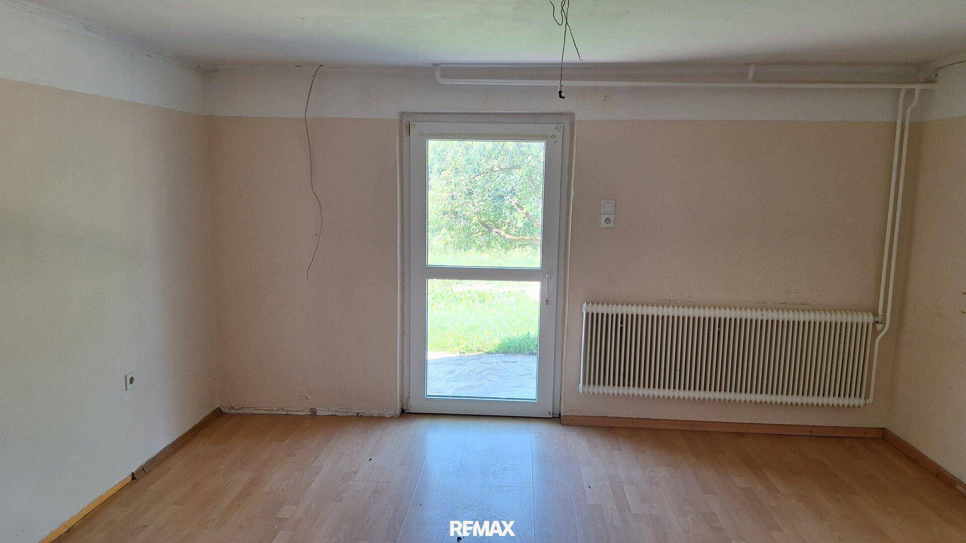 Leeres Zimmer mit Holzboden, Heizkörper und Tür zum Gartenbereich.
