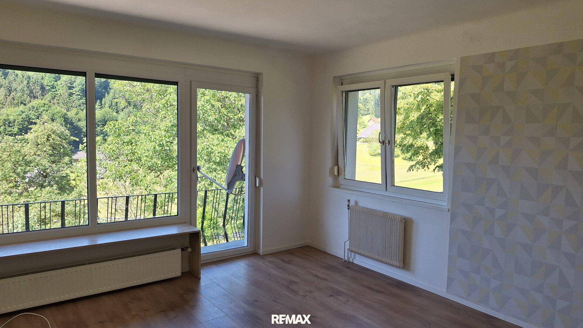 Helles Zimmer mit Holzboden, großen Fenstern und Zugang zu einem Balkon mit Waldblick.