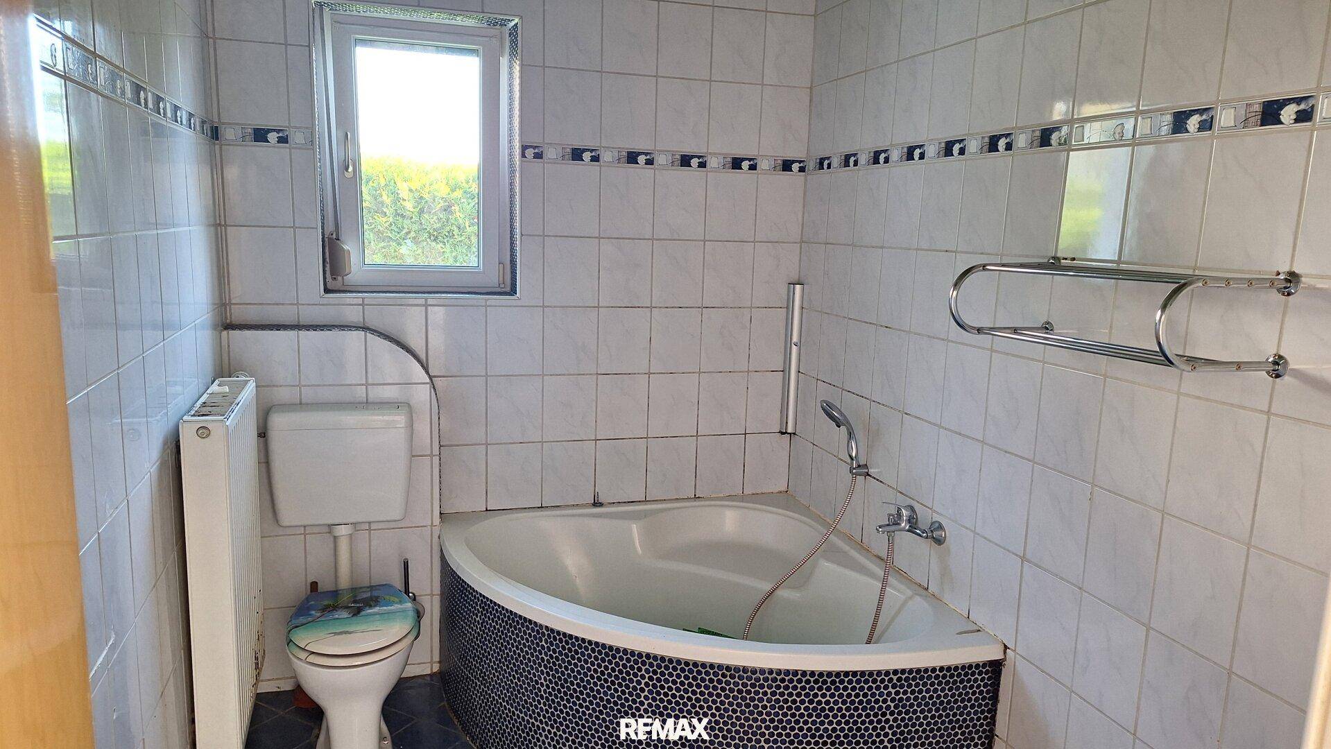 Badezimmer mit Eckbadewanne, weißen Fliesen und einem Fenster für Tageslicht.