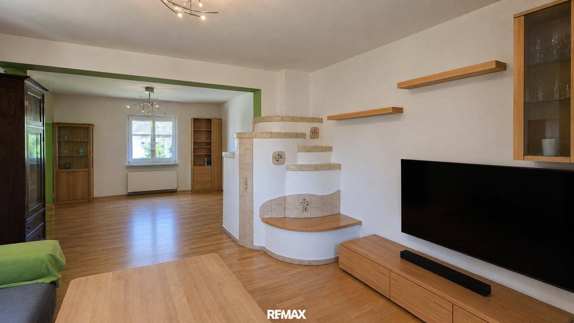 Helles Wohnzimmer mit Holzboden, modernem Kachelofen und Blick in einen weiteren Raum.