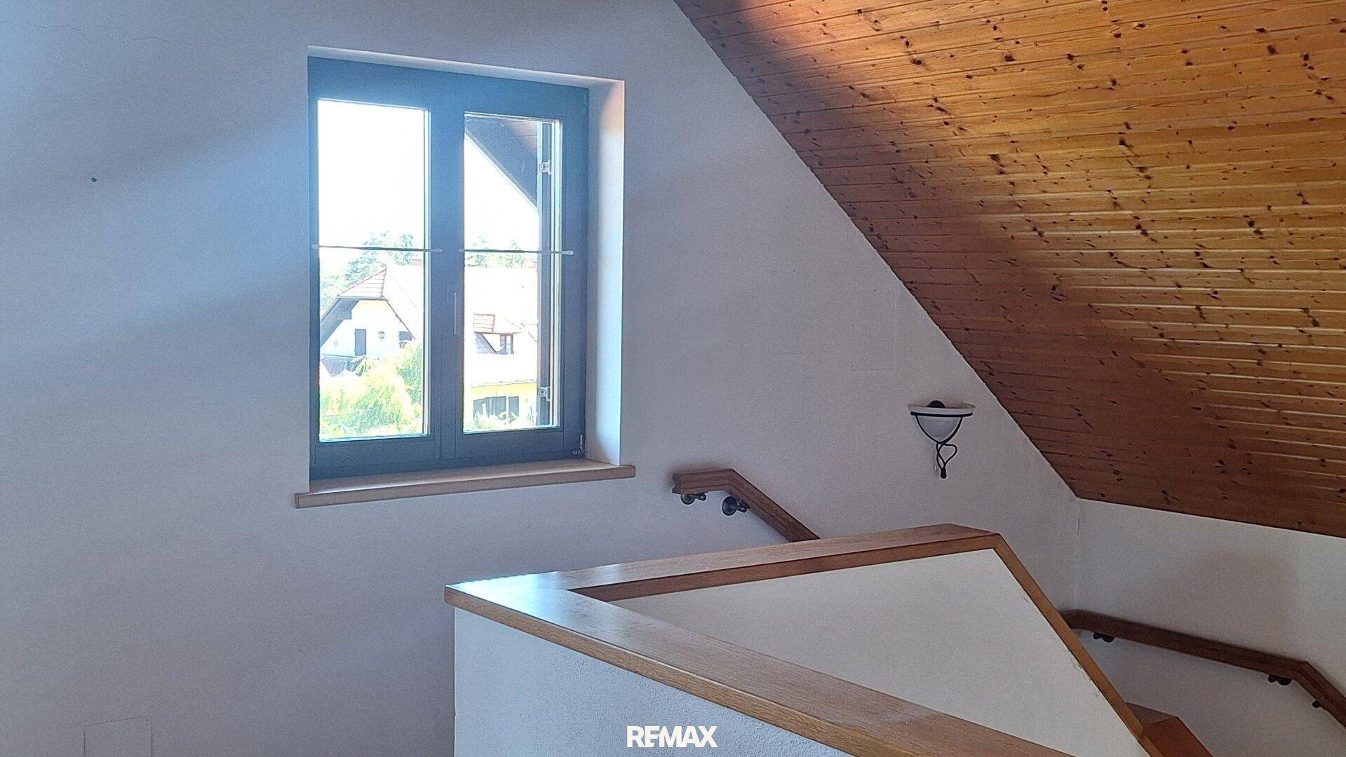 Heller Treppenaufgang mit Holzgeländer, Fenster und einer Holzdecke unter einer Dachschräge.