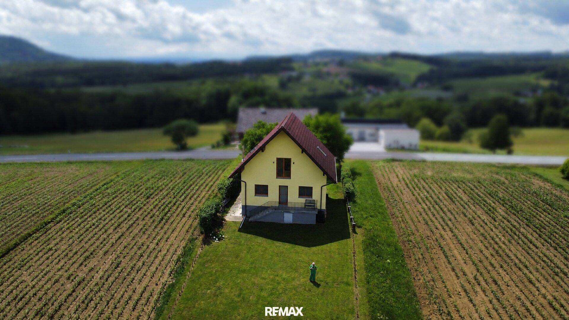 Luftaufnahme eines gelben Einfamilienhauses umgeben von Feldern und grüner Landschaft unter bewölktem Himmel.