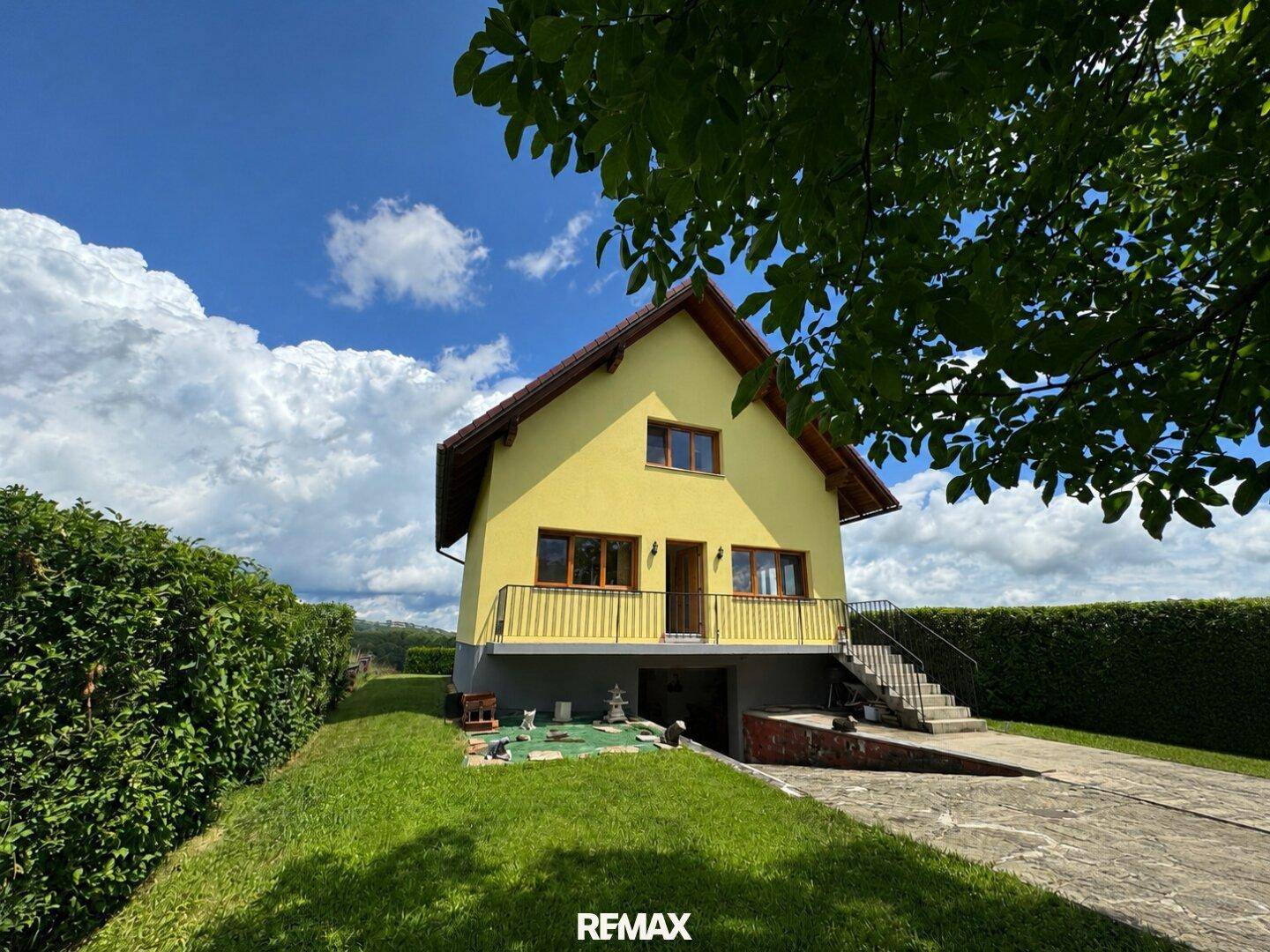 Gelbes Einfamilienhaus mit rotem Dach, Balkon und Außentreppe, umgeben von grüner Hecke und Rasen.