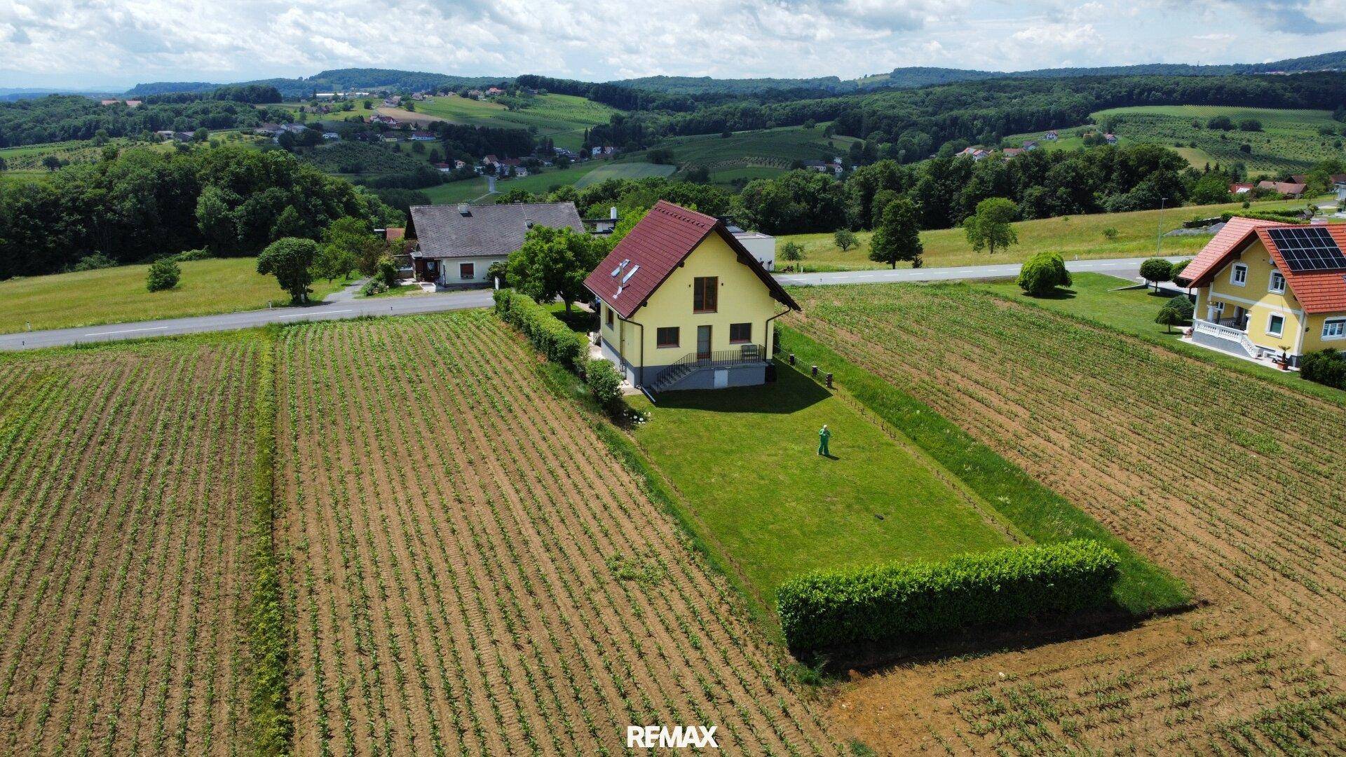 Vogelperspektive eines gelben Hauses mit rotem Dach, umgeben von Feldern und einer weiten grünen Landschaft.