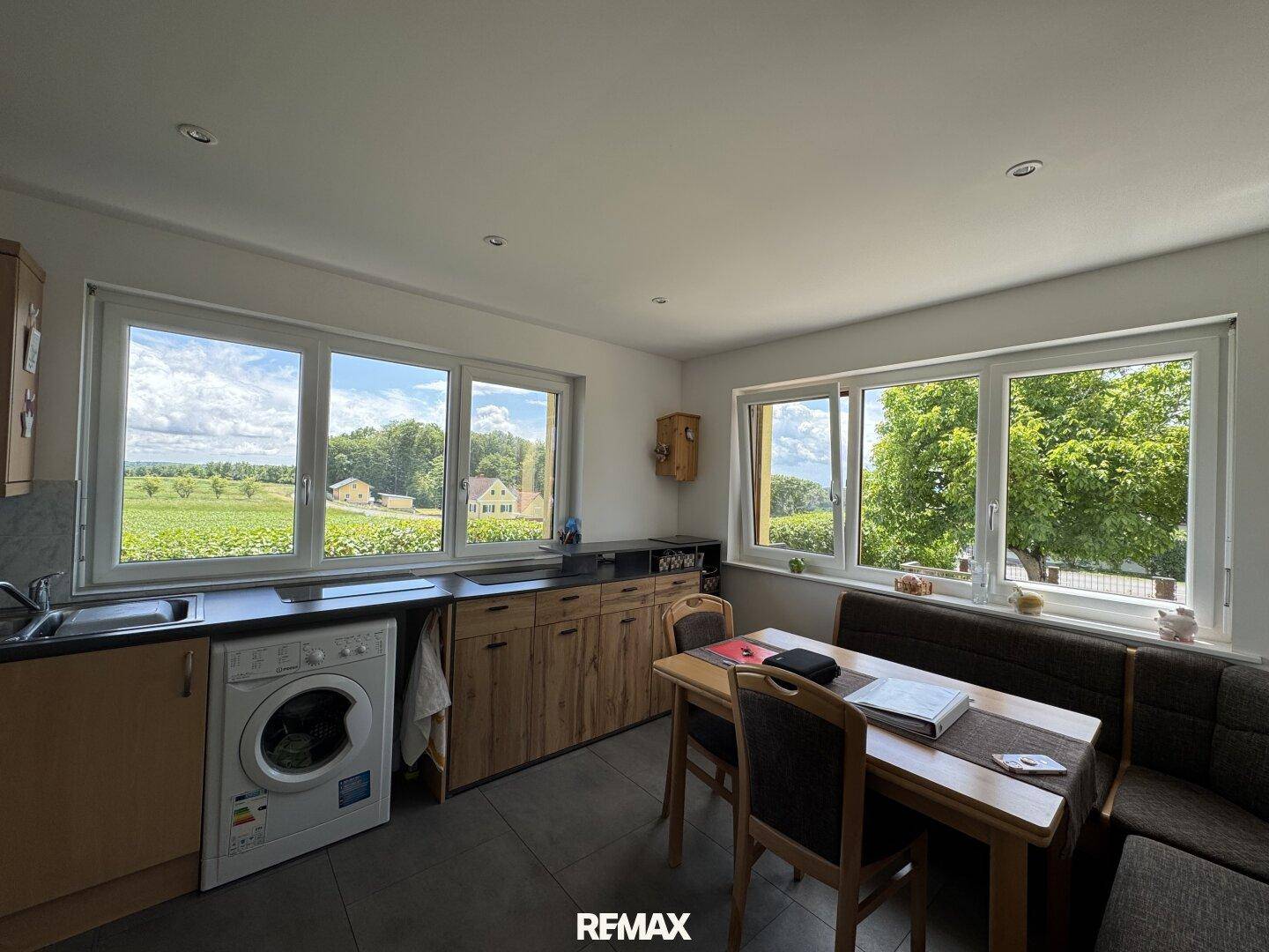 Helle Küche mit Holzelementen, Essbereich und weitem Blick auf die grüne Landschaft durch große Fenster.