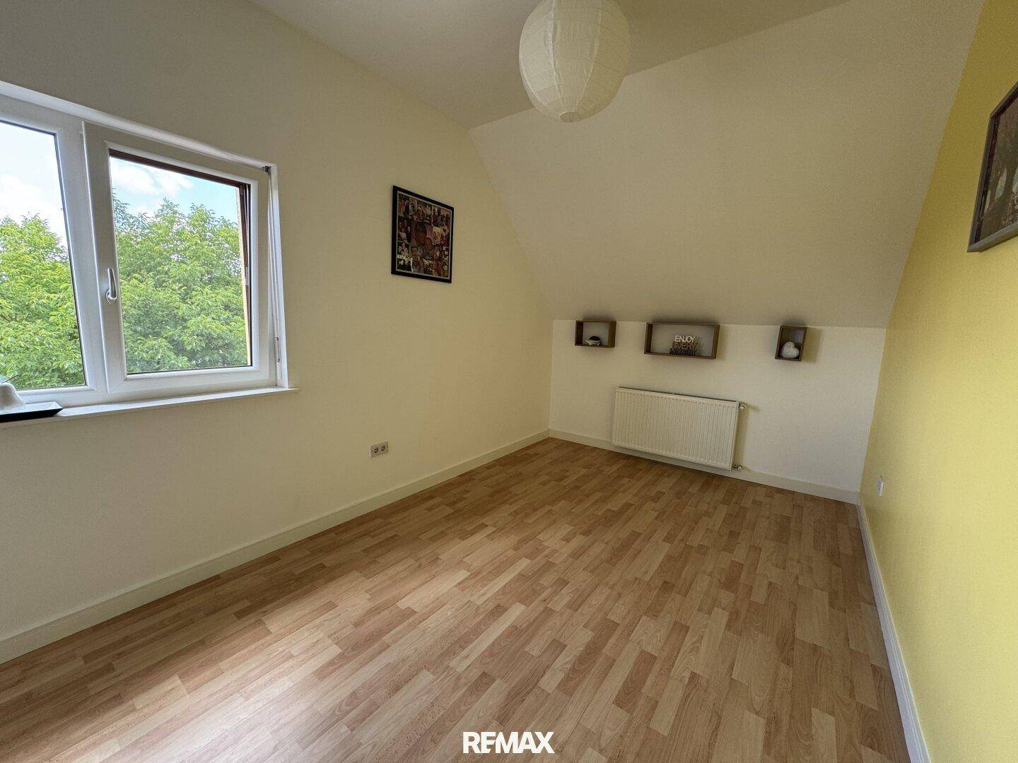 Helles Schlafzimmer mit Laminatboden, Dachschräge und einem Fenster mit Blick ins Grüne.