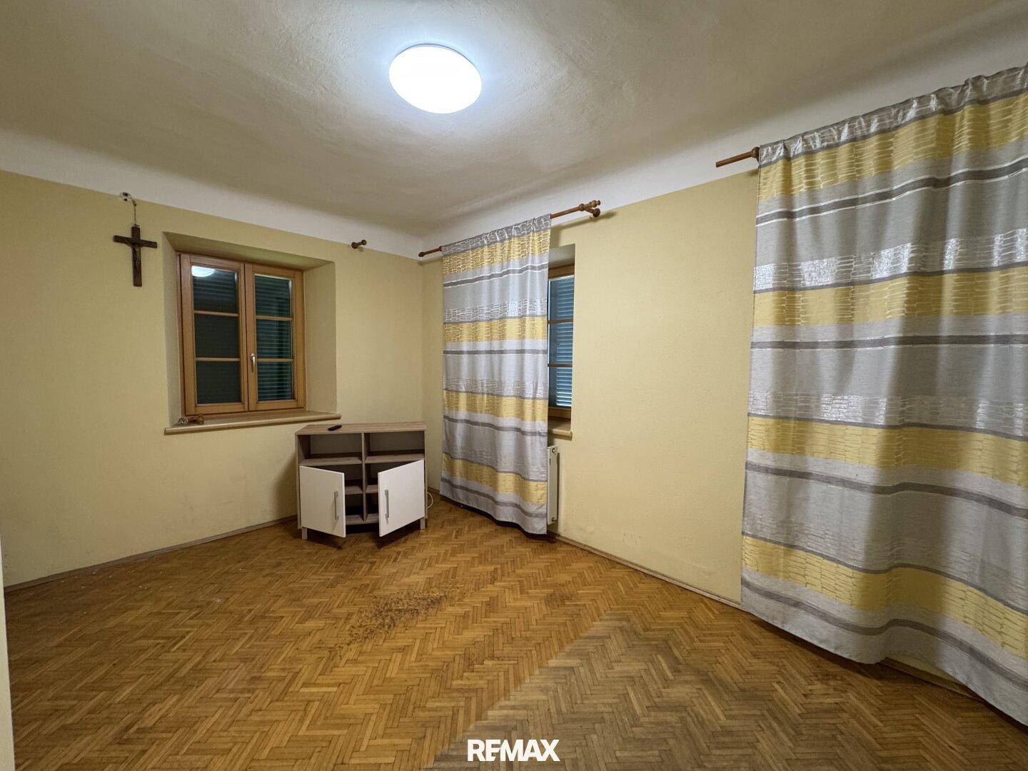Helles Zimmer mit Fischgrätparkett, gelben Wänden und zwei Fenstern mit gestreiften Vorhängen.