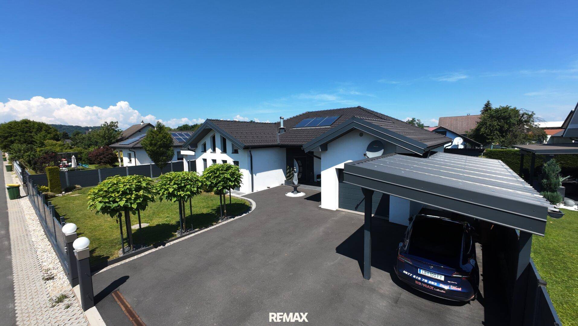 Weitläufige Luftaufnahme des modernen Hauses mit Carport, Einfahrt und gepflegtem Vorgarten unter strahlendem Himmel.