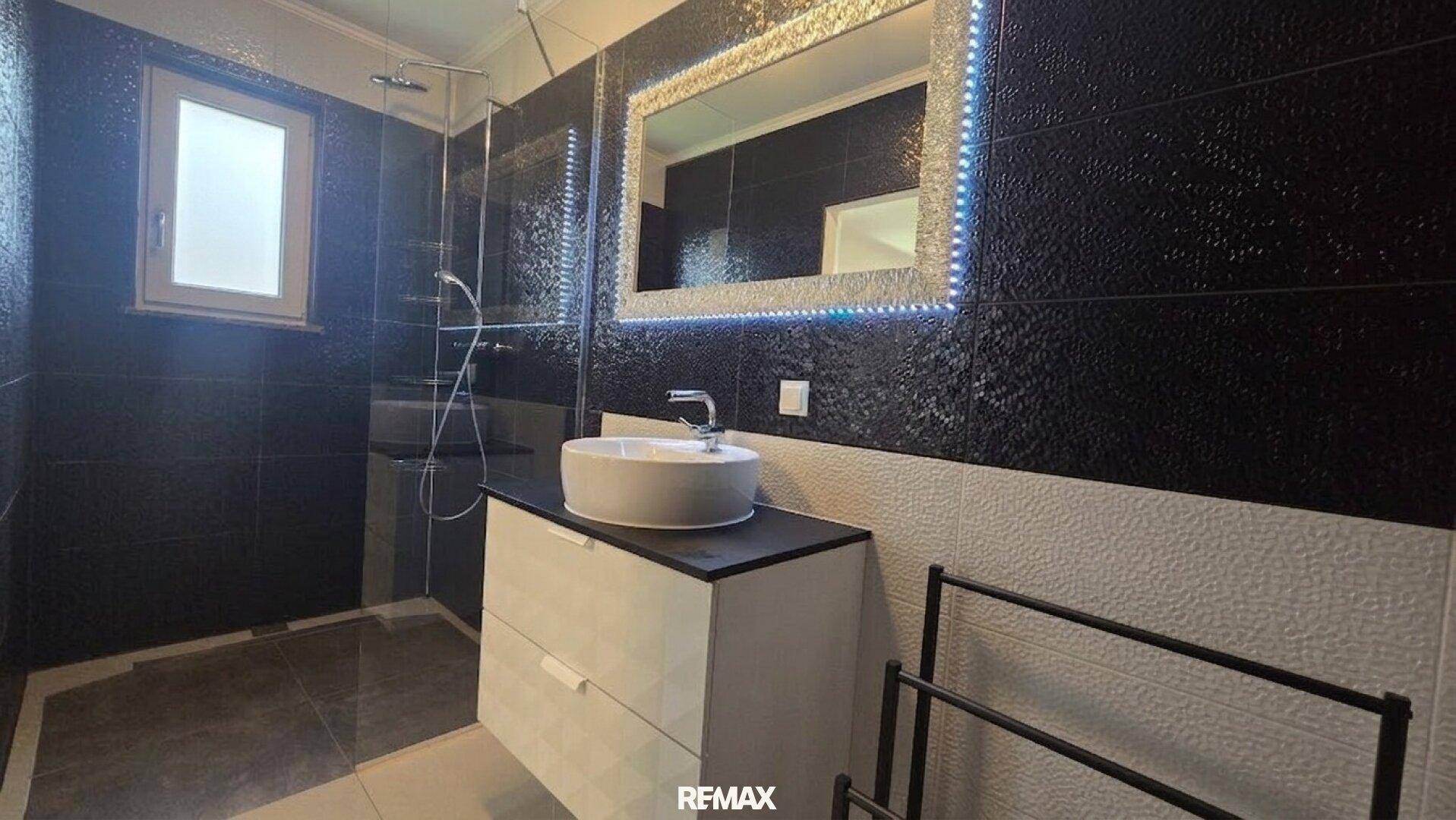 Elegantes Badezimmer mit begehbarer Dusche, beleuchtetem Spiegel und dunklen Fliesen.