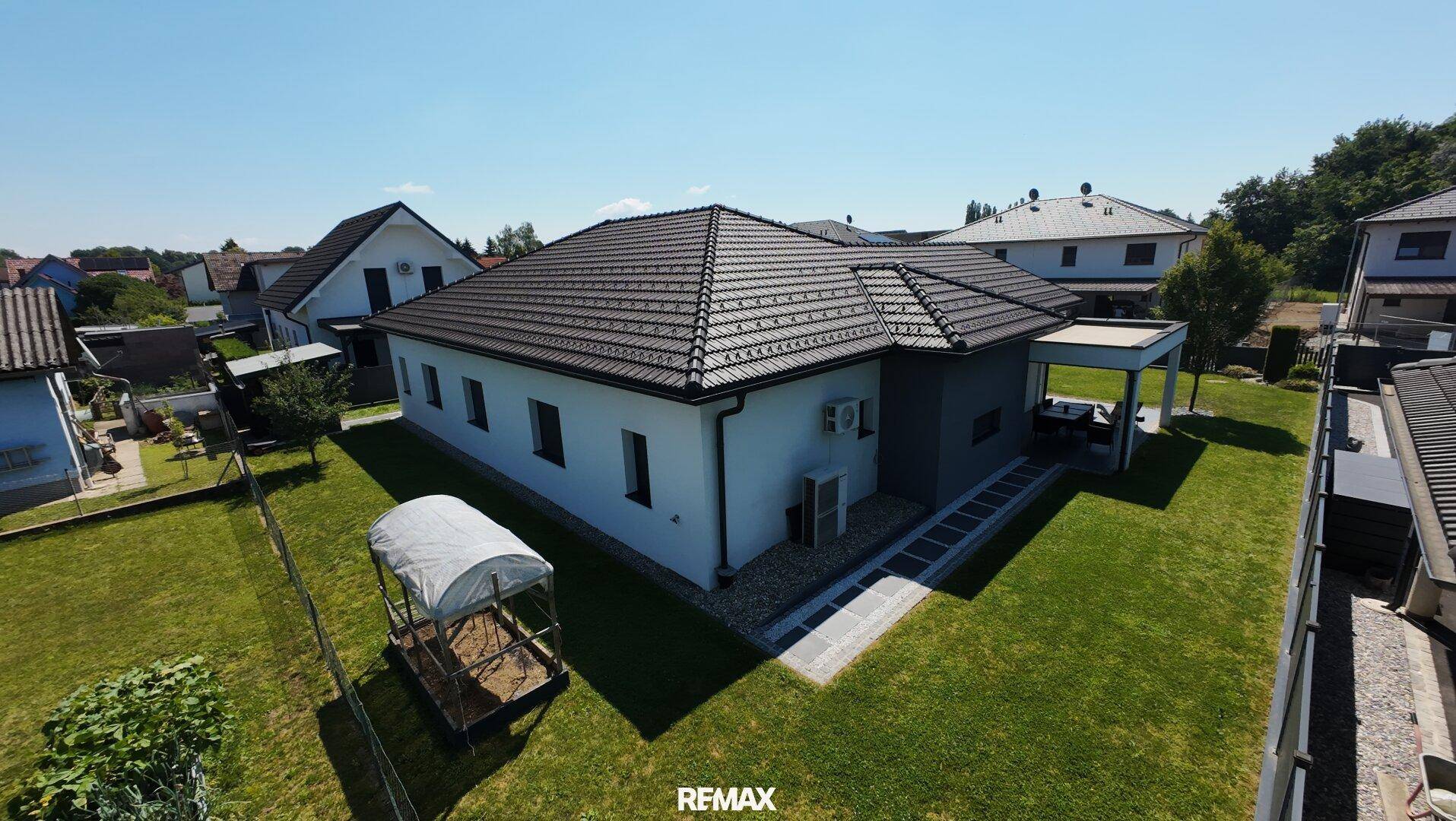 Rückansicht des Hauses mit großem Garten, Terrasse und einem kleinen Gewächshaus unter blauem Himmel.