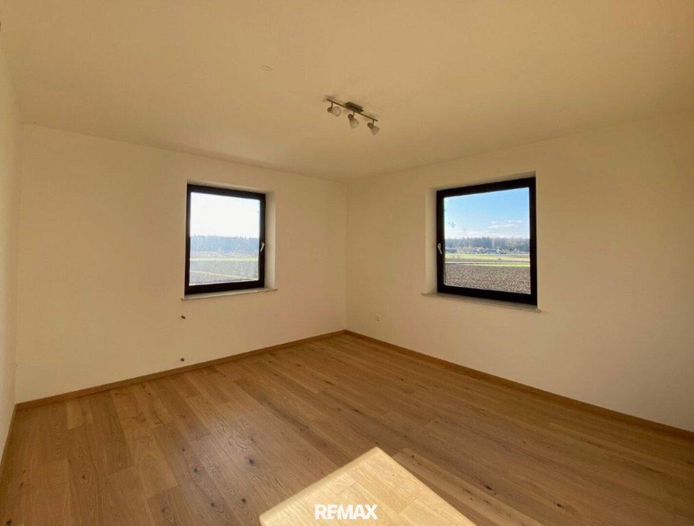 Helles Schlafzimmer mit Holzboden und zwei Fenstern mit Blick auf die Felder.