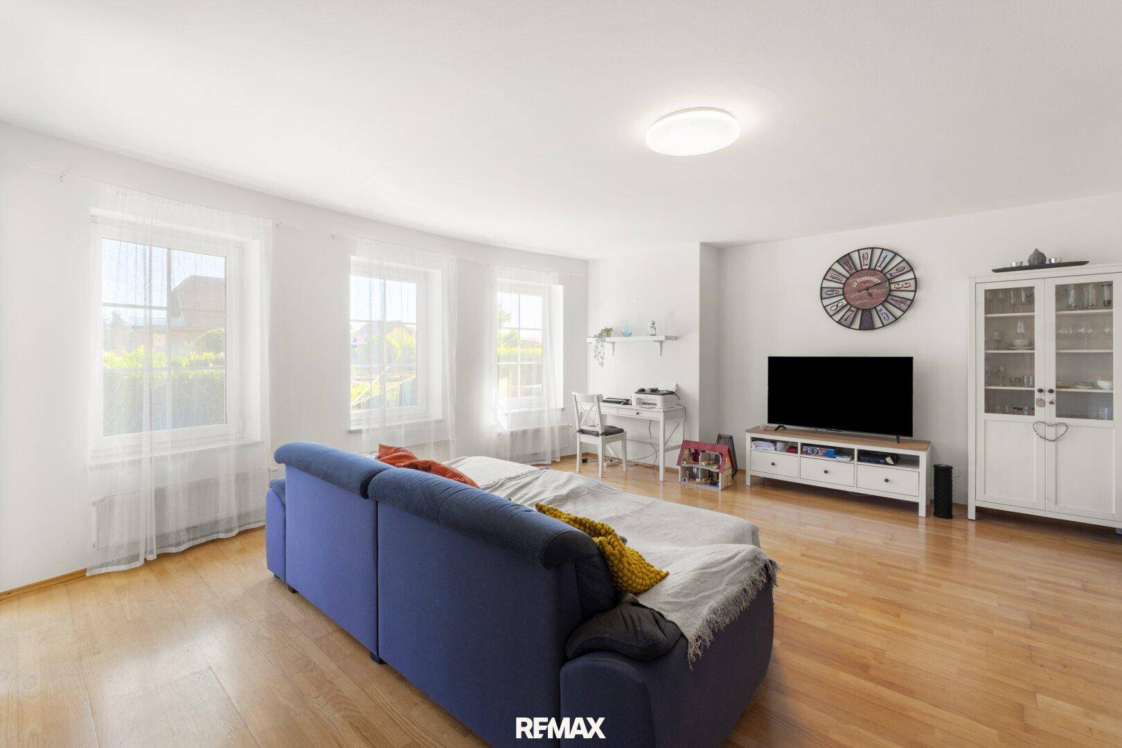 Geräumiges Wohnzimmer mit hellem Holzboden, blauer Couch und großen Fenstern für viel Tageslicht.