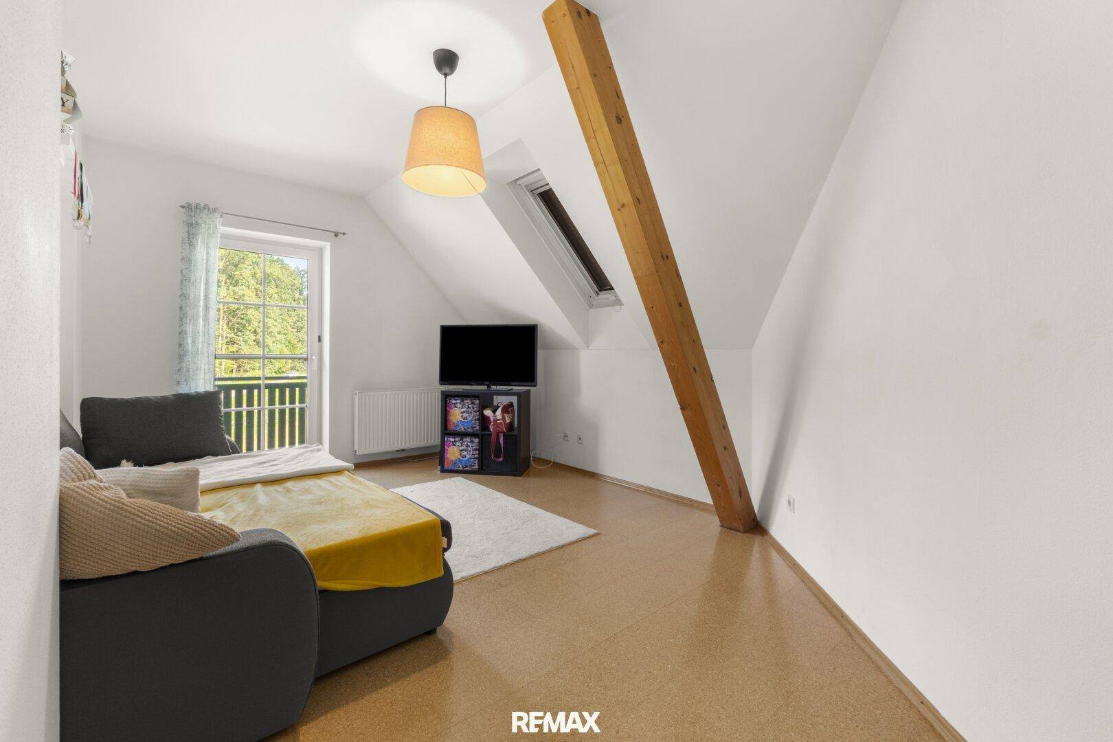 Helles Dachzimmer mit Sofa, sichtbarem Holzbalken, Balkonzugang und Korkboden.